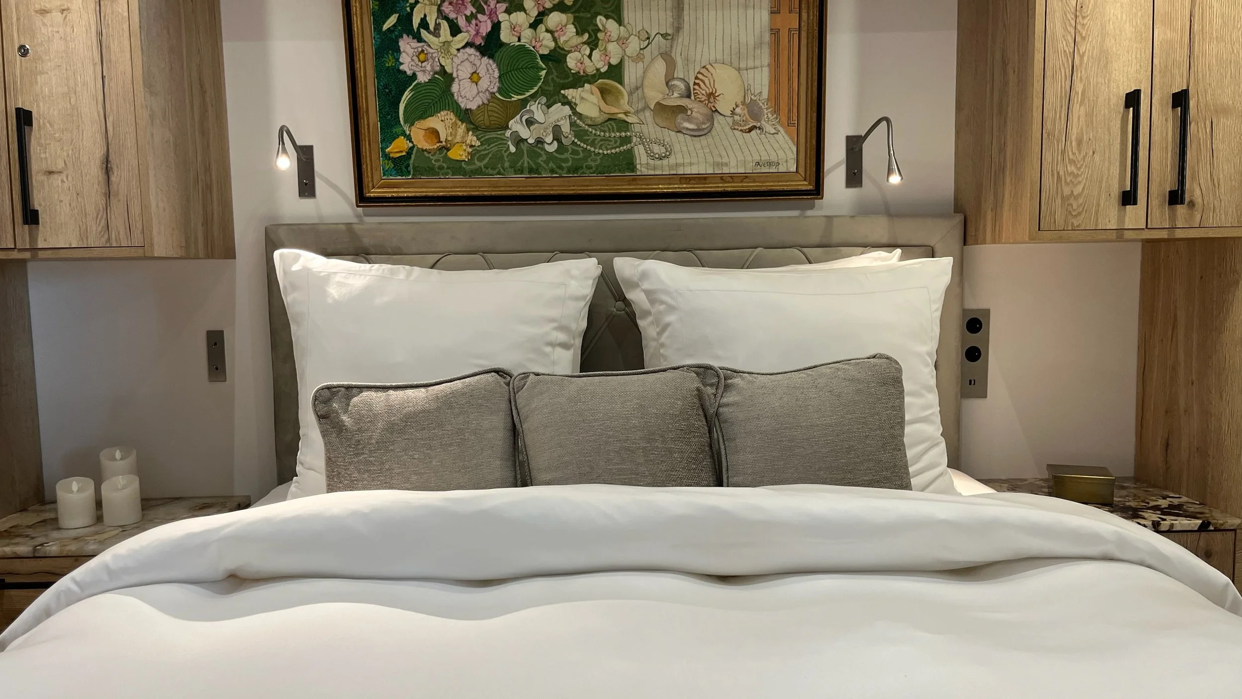 A neatly made bed featuring two large white pillows, two smaller gray cushions in front, and a beige upholstered headboard. On either side of the bed, there are small wooden nightstands with candles and other small items, with wall-mounted reading li