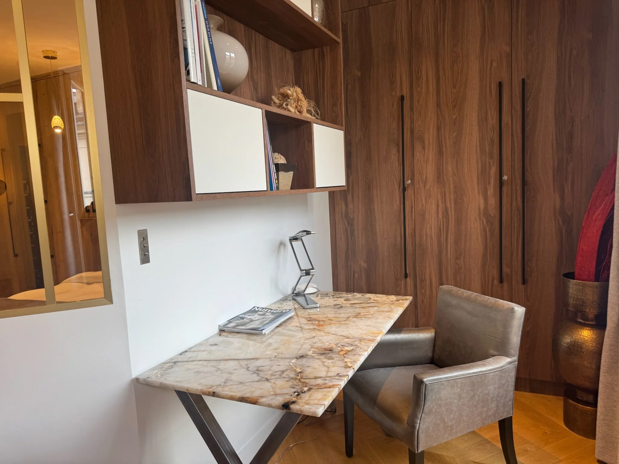 A corner of a room with a marble top desk, a beige armchair, and a wooden wall with black handles on built-in cabinets. A bookshelf with decorative items and books is mounted above the desk.