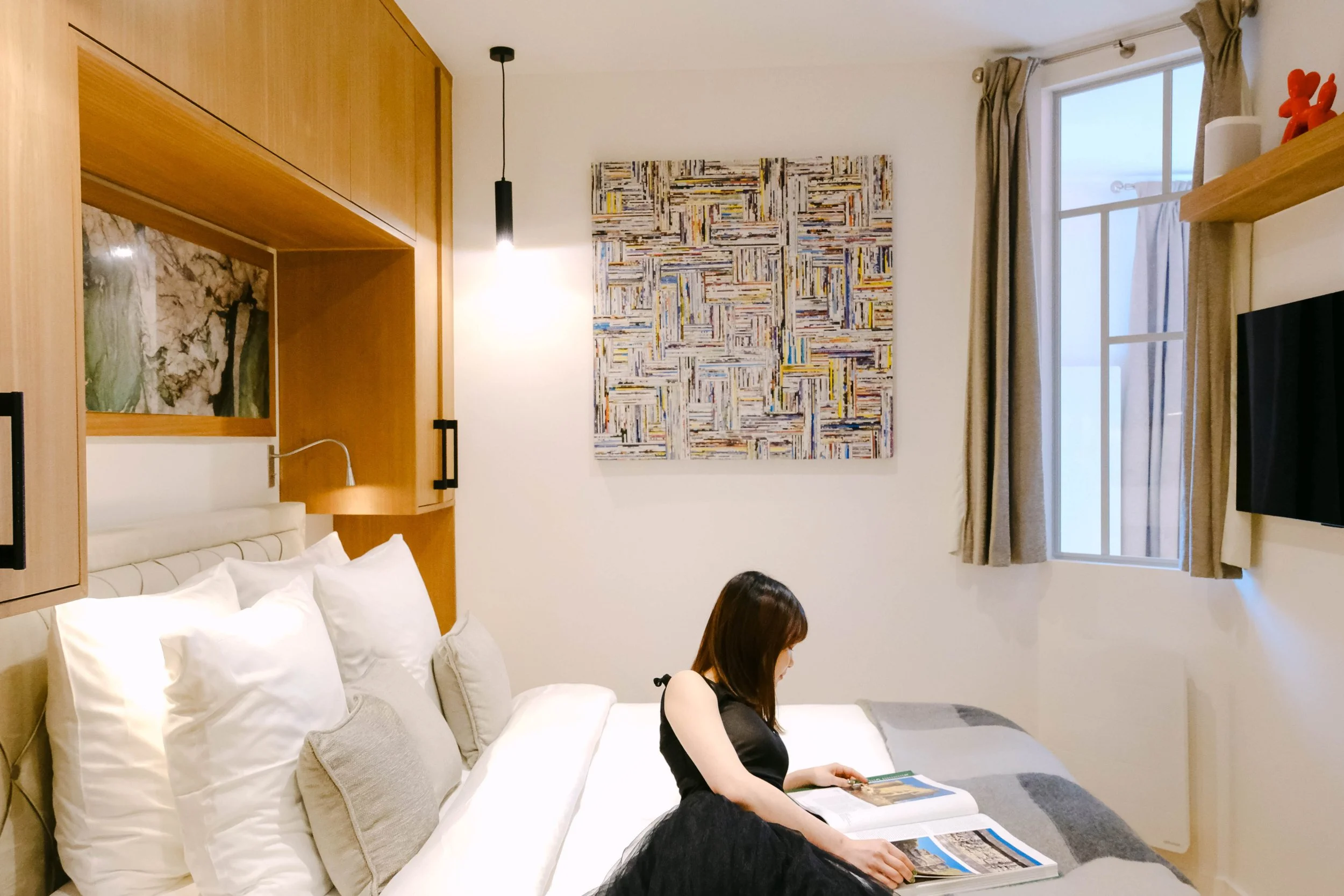 A woman sitting on a bed in a modern hotel room, looking at magazines. The room has a white wall with colorful abstract artwork, a window with curtains, a flat-screen TV, and wooden furniture.