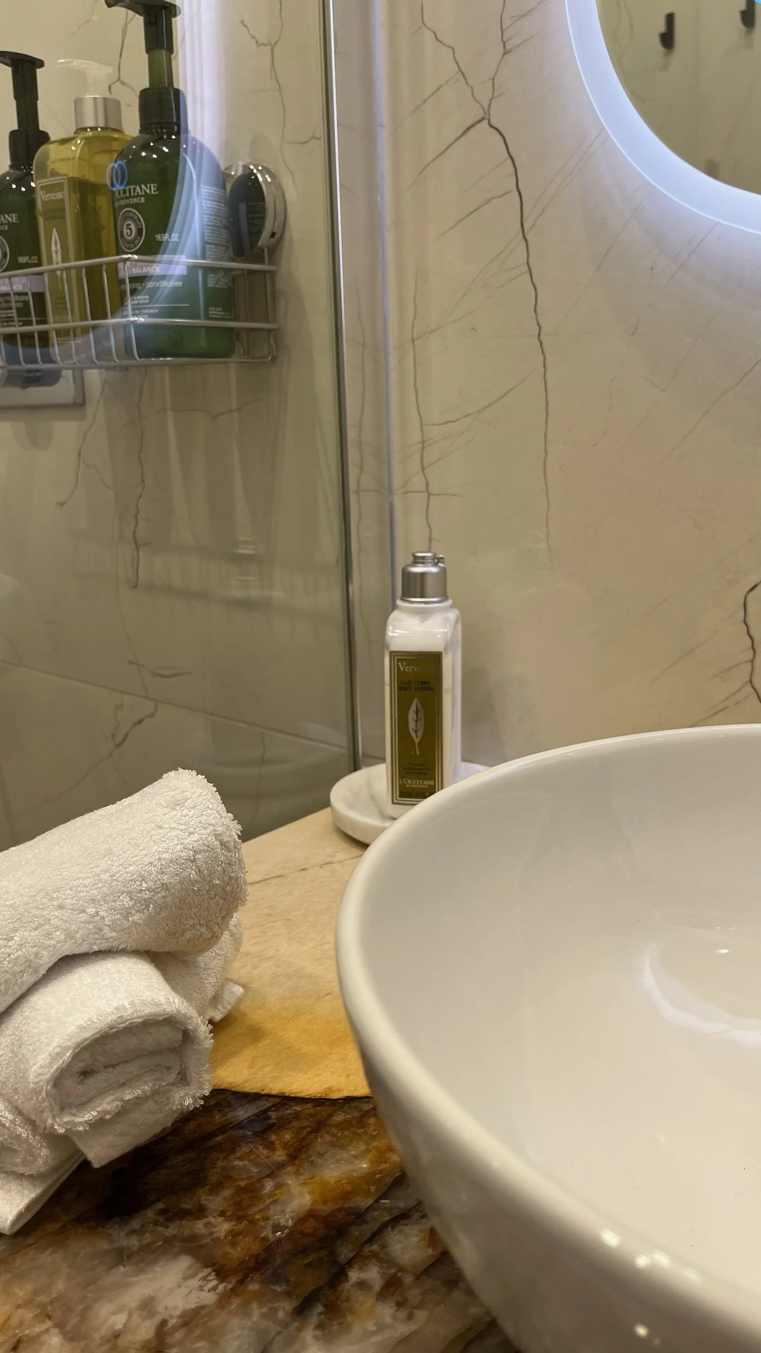 A bathroom countertop with a white towel, a black marble surface, a white sink, a bottle of perfume labeled "Vero" on a round tray, a round mirror with LED lighting, and a shower area with a shelf holding various bottles of shampoo and conditioner.