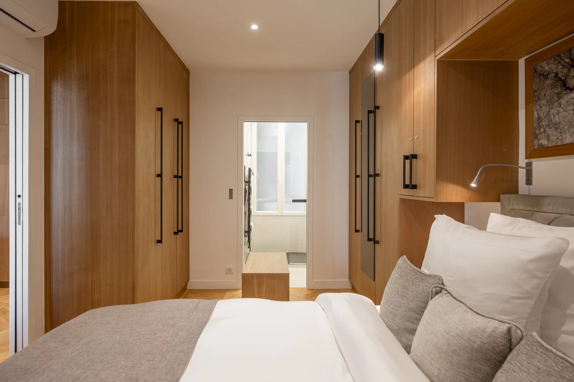Modern bedroom with wood paneling, a bed with white and gray linens, wall-mounted TV, abstract art, and large window with curtains.