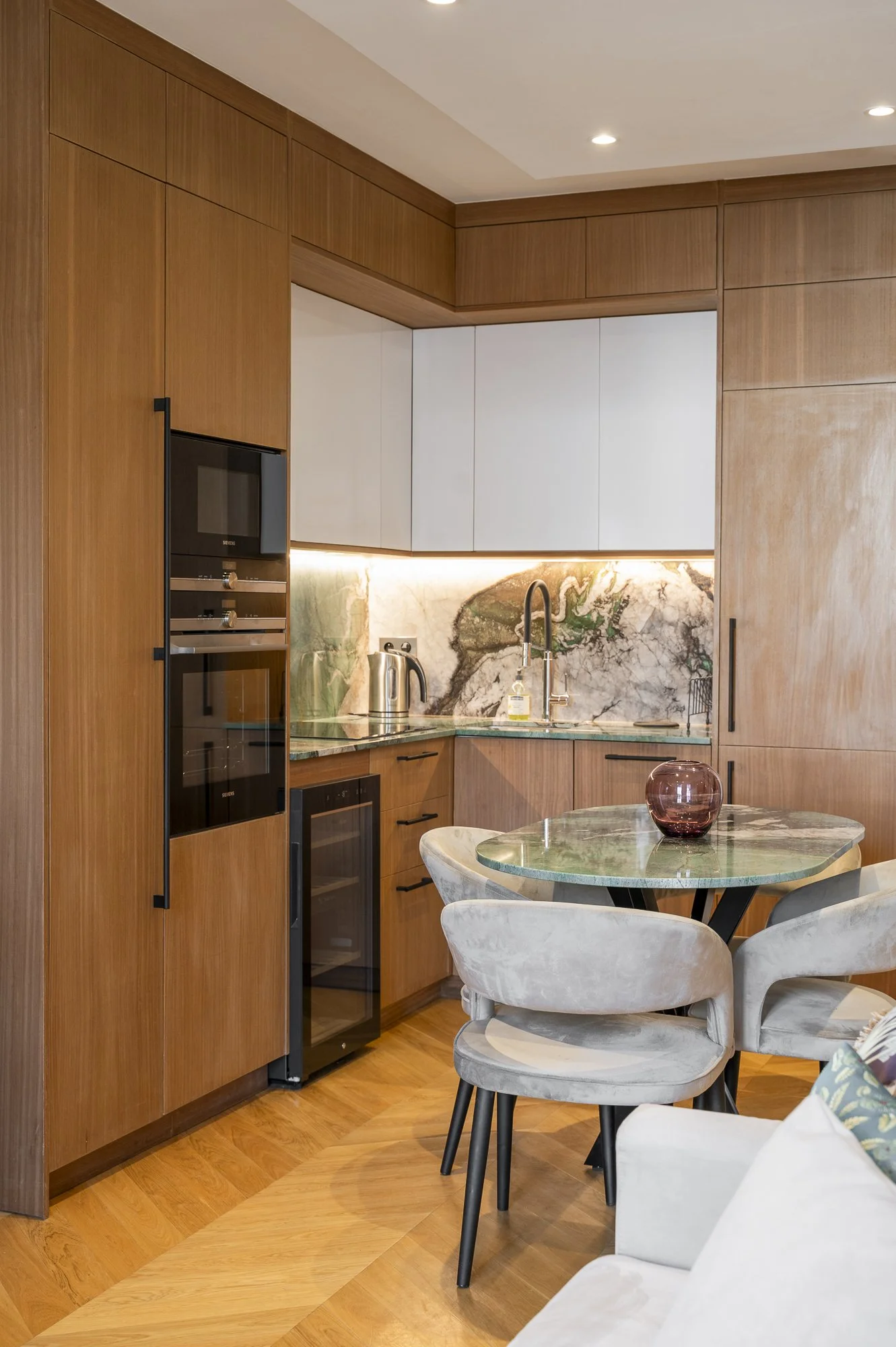 Modern kitchen with wooden cabinetry, marble backsplash, and a small dining table with four velvet chairs.