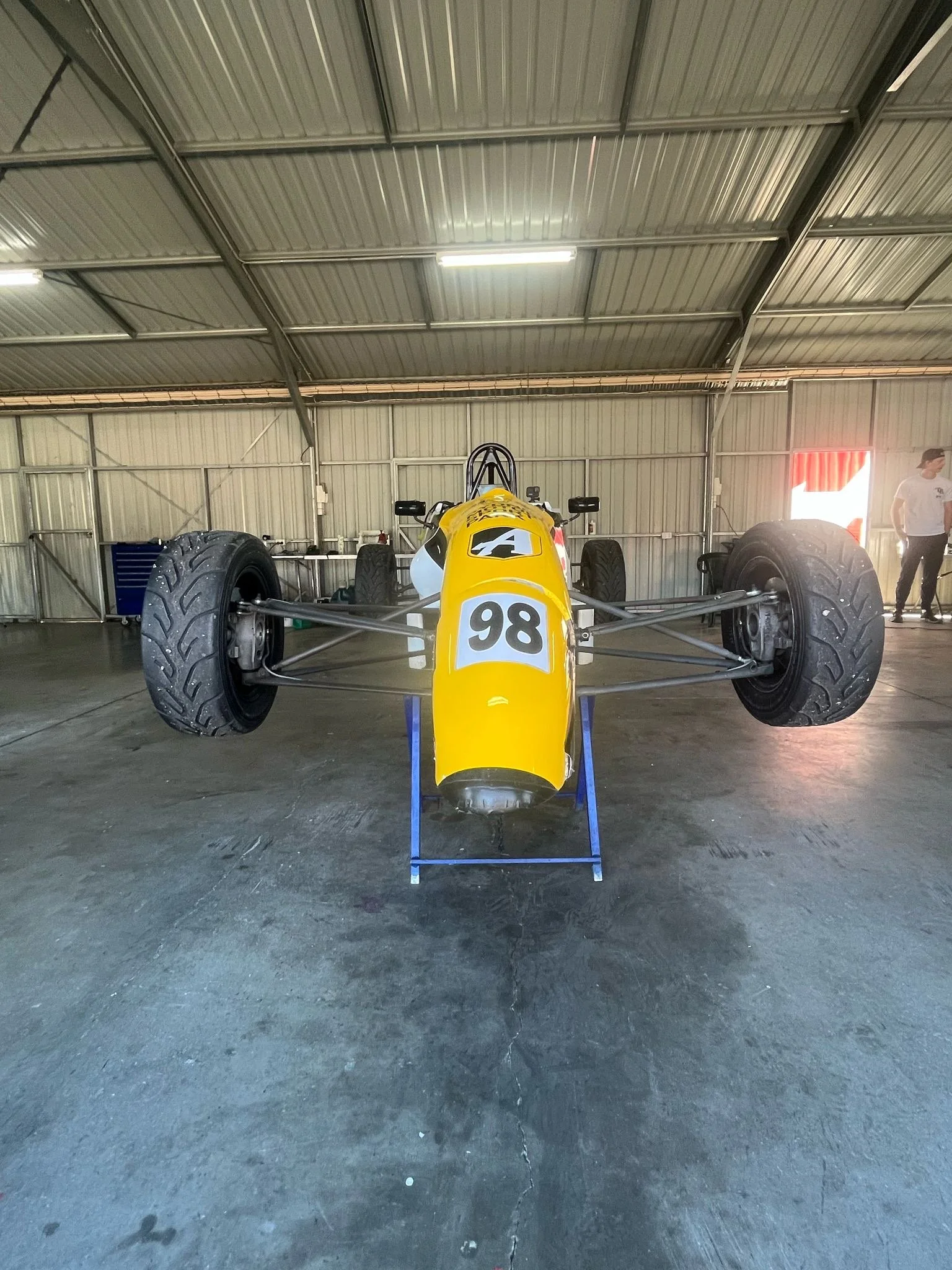 A vintage open-wheel yellow race car with the number 98 on the front, inside a garage or workshop with a metal roof and concrete floor.