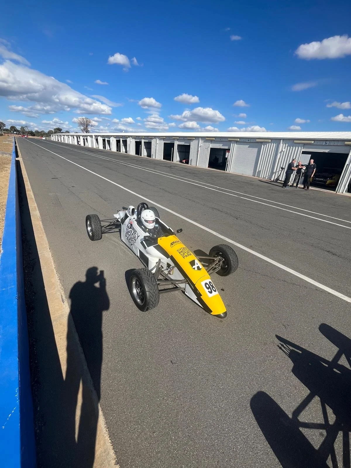 A vintage race car with the number 98 on the front, parked on a race track pit lane, with garage units and two men talking in the background, under a partly cloudy sky.