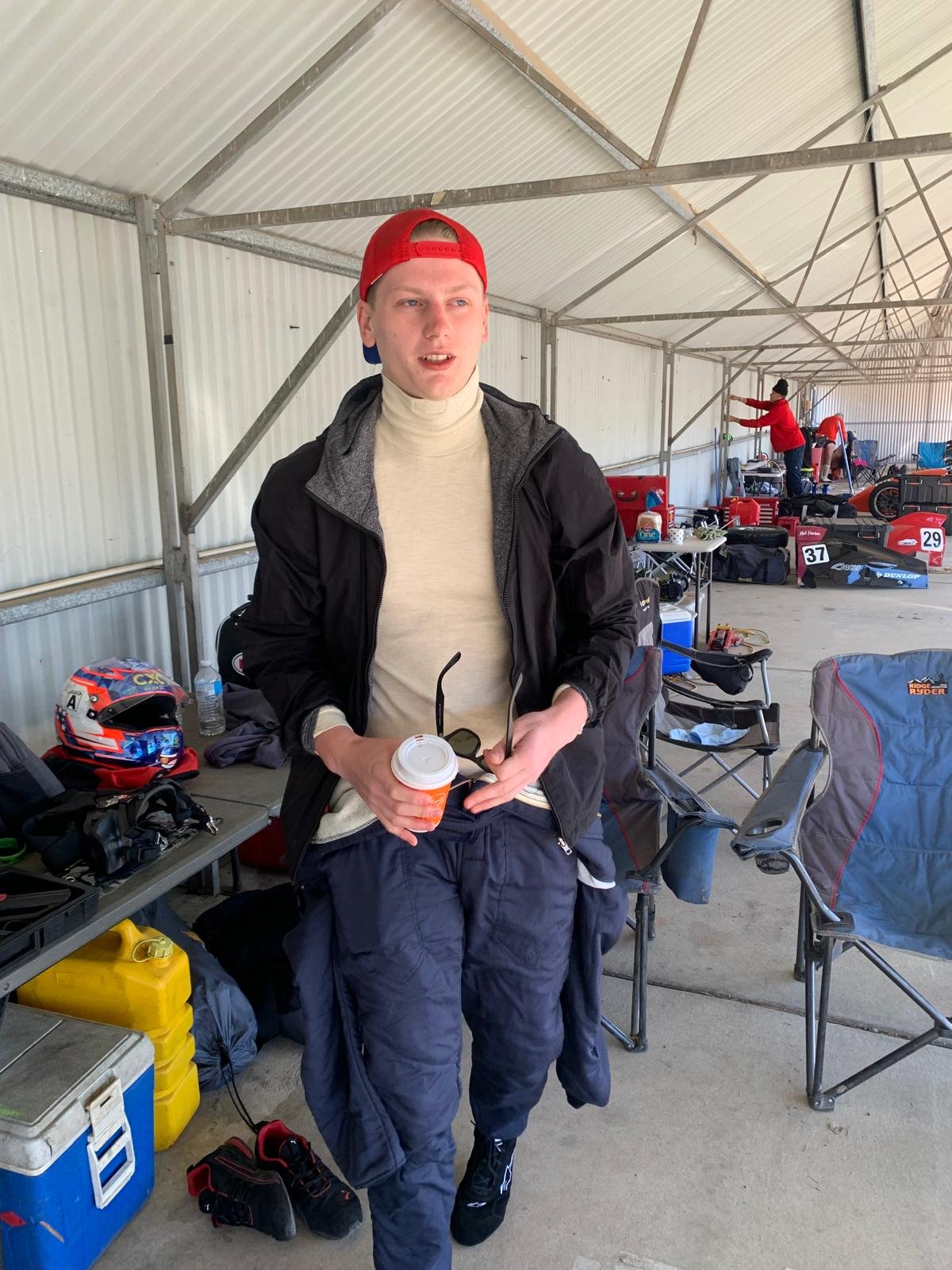 Young man wearing racing gear, standing in a garage area with racing helmets, tools, and equipment around him.