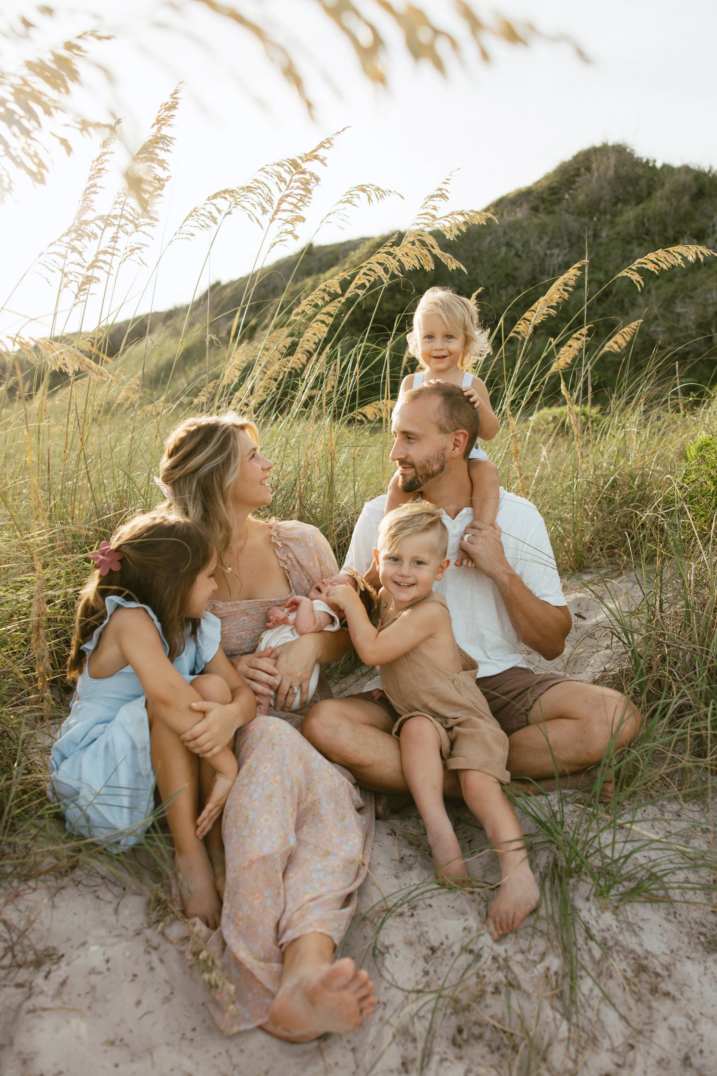 A family of six sitting on the sand at the beach surrounded by tall grass, with a hill in the background, enjoying a pleasant moment during sunset.