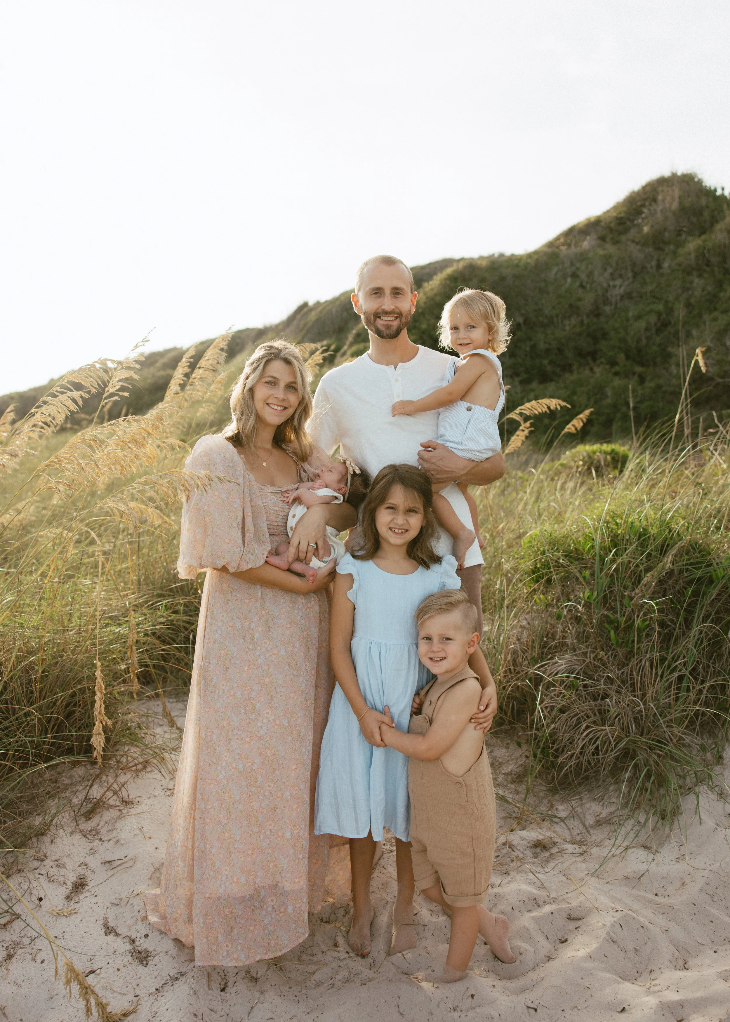 A family of seven standing on a sandy beach with tall grasses and green hills in the background, smiling and embracing each other during sunset.
