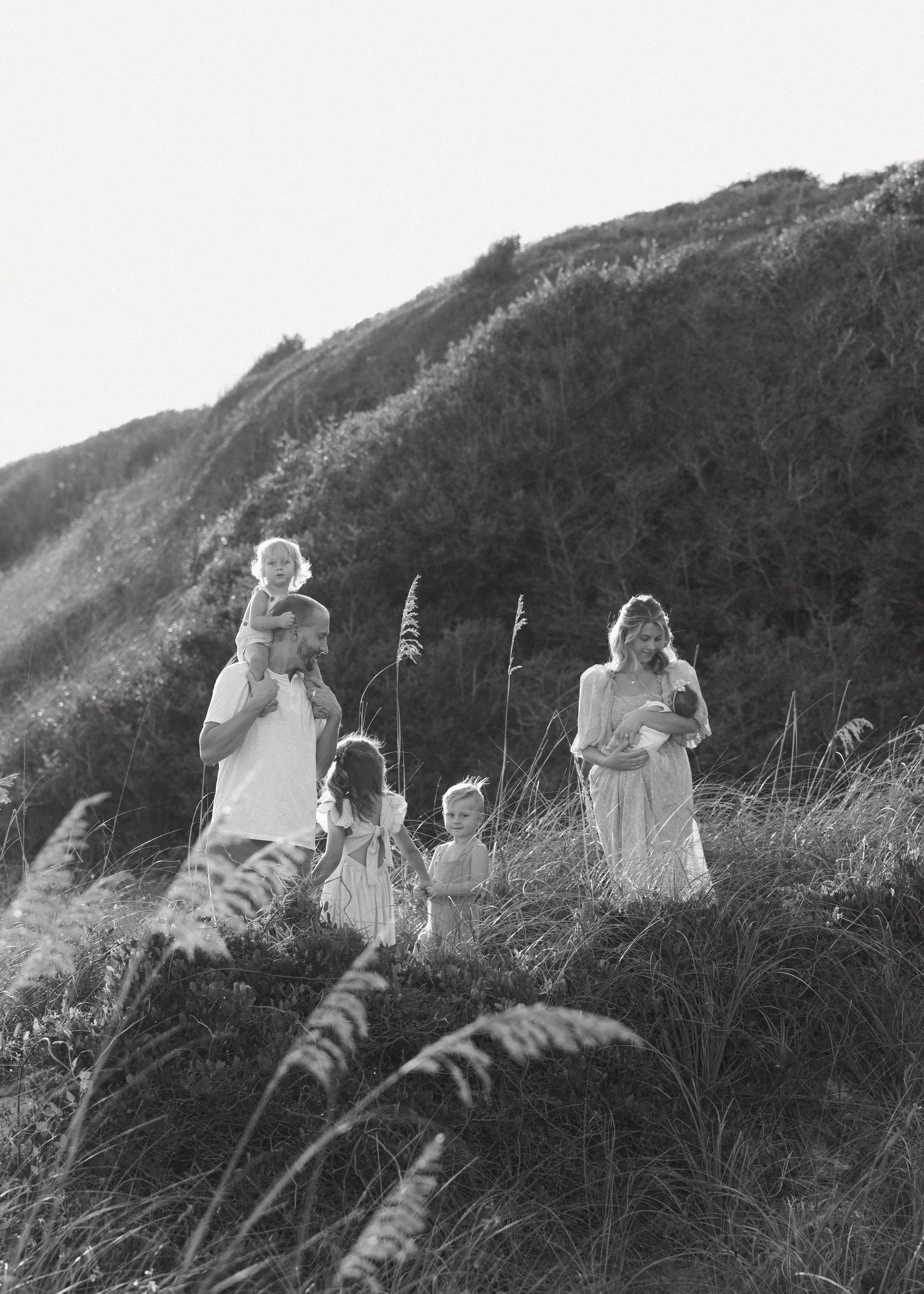 A family outdoors walking through tall grass on a hillside, with a man carrying a child on his shoulders and a woman holding a baby, three young children holding hands, and a hilly landscape in the background.
