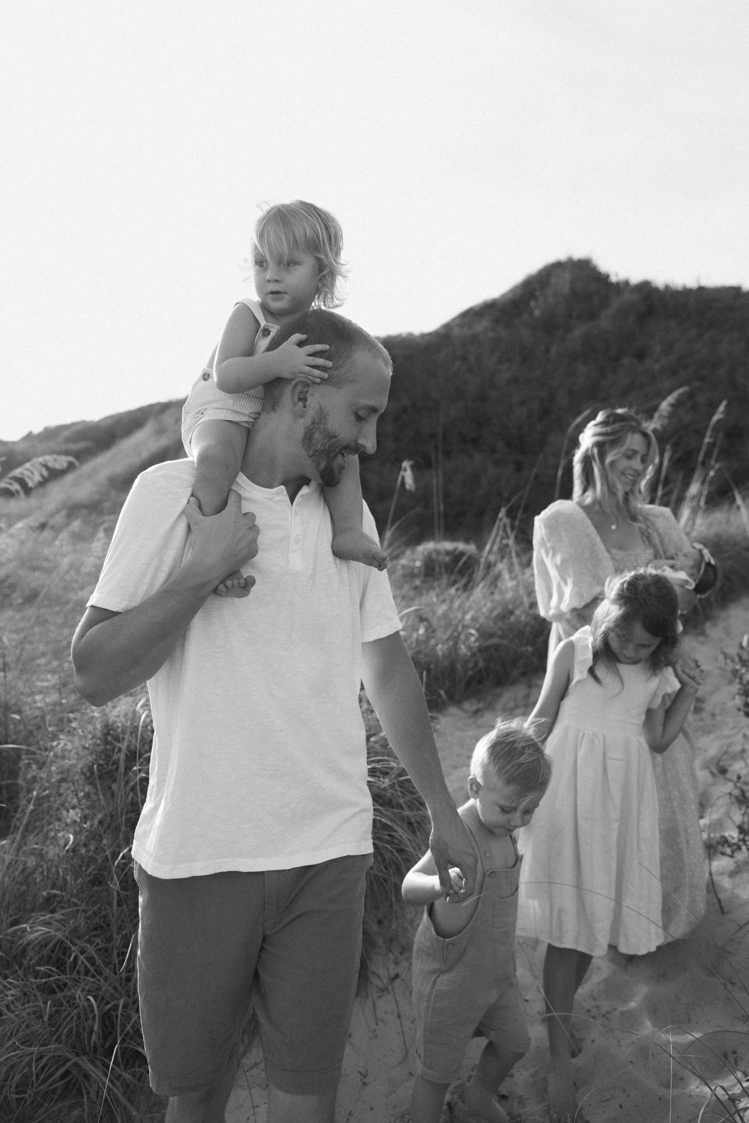 A father walking on a sandy trail with his children, holding hands and carrying one on his shoulders, in a natural outdoor setting.