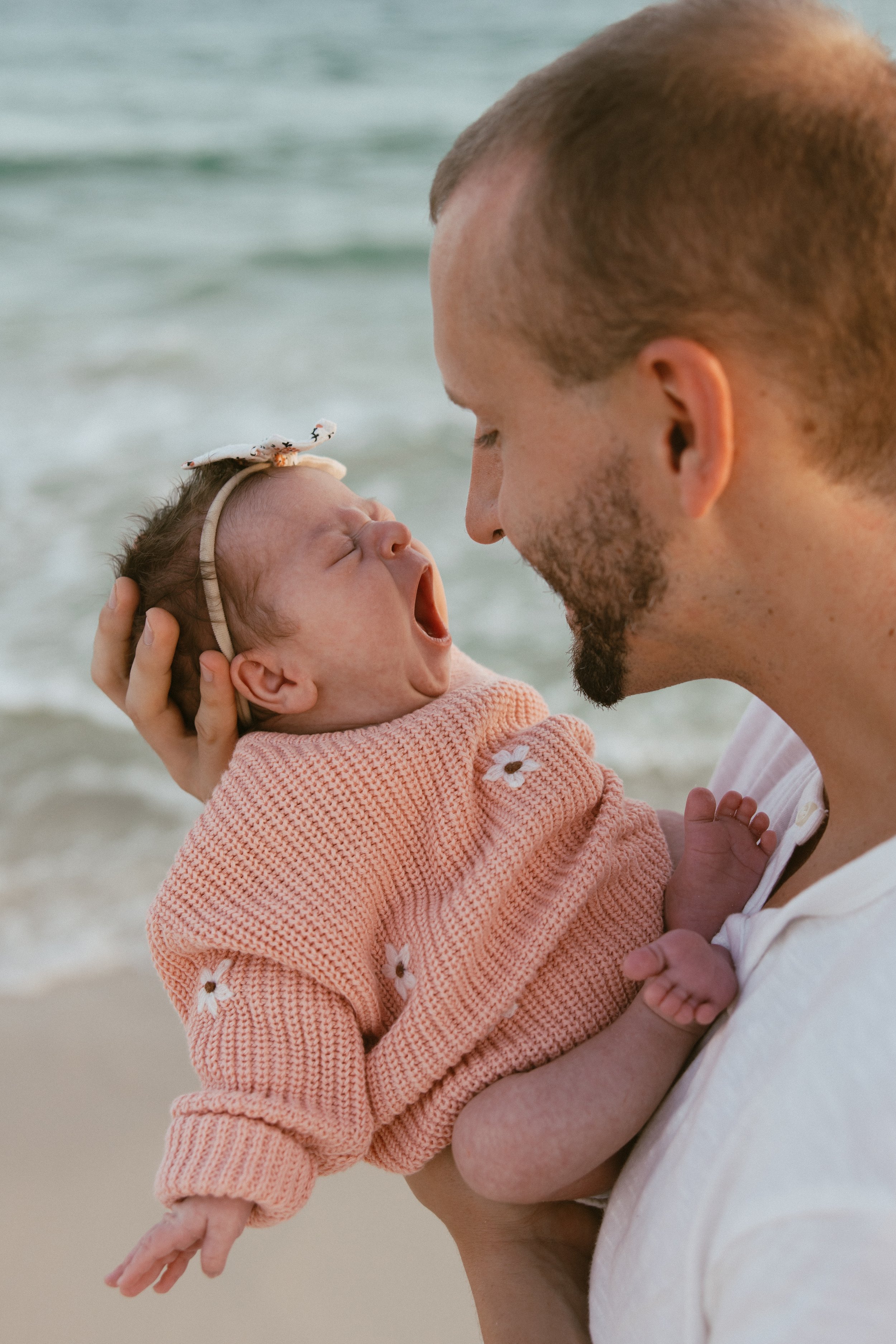 A man holding a yawning baby girl on a beach, with ocean waves in the background.
