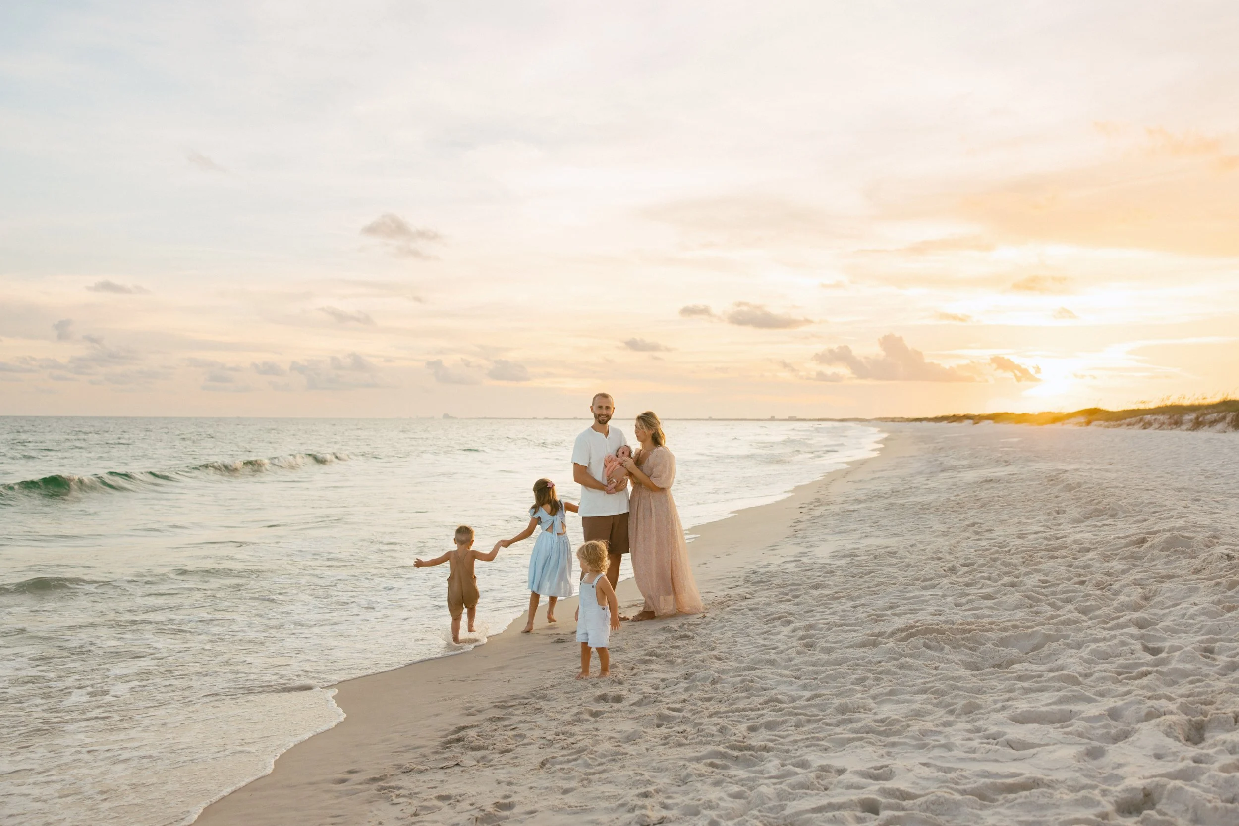 A family of six enjoying their time on the beach at sunset, with two adults, three children, and a baby, all dressed in light summer clothes near the water.