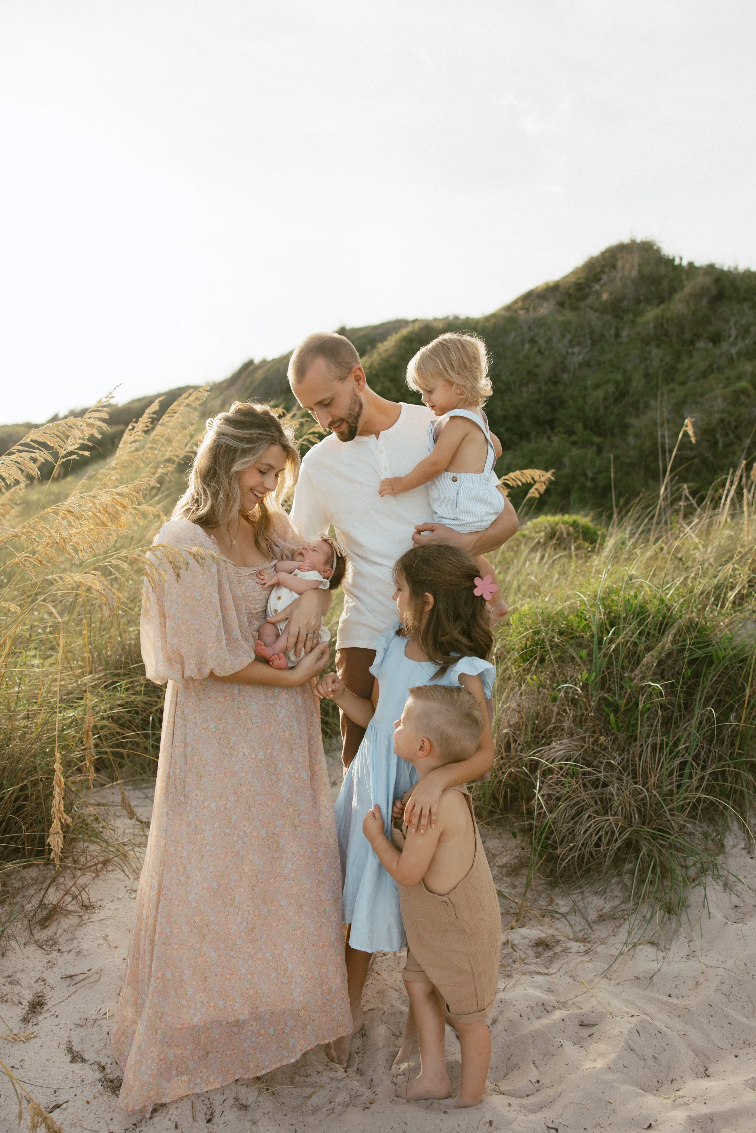 Family with two adults and five children standing on a beach during sunset, with grassy dunes and hills in the background.