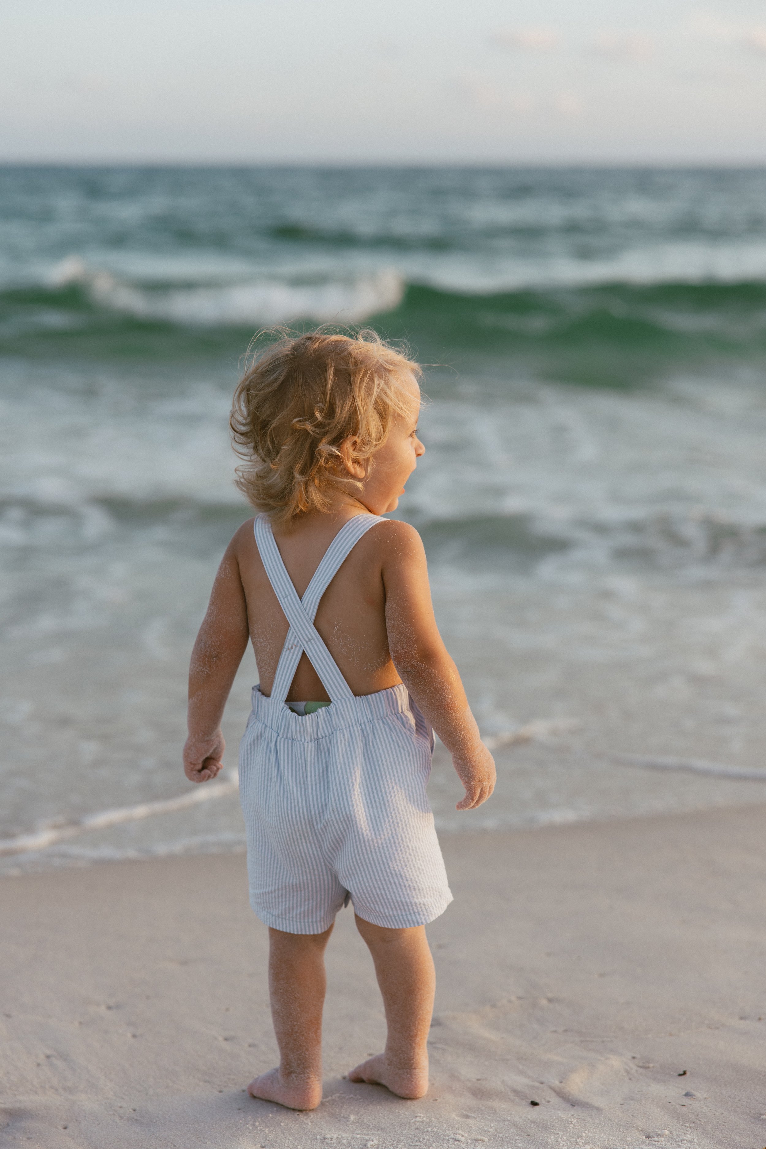 A young child with curly blonde hair standing barefoot on the beach, facing the ocean, wearing white and light blue striped overalls.