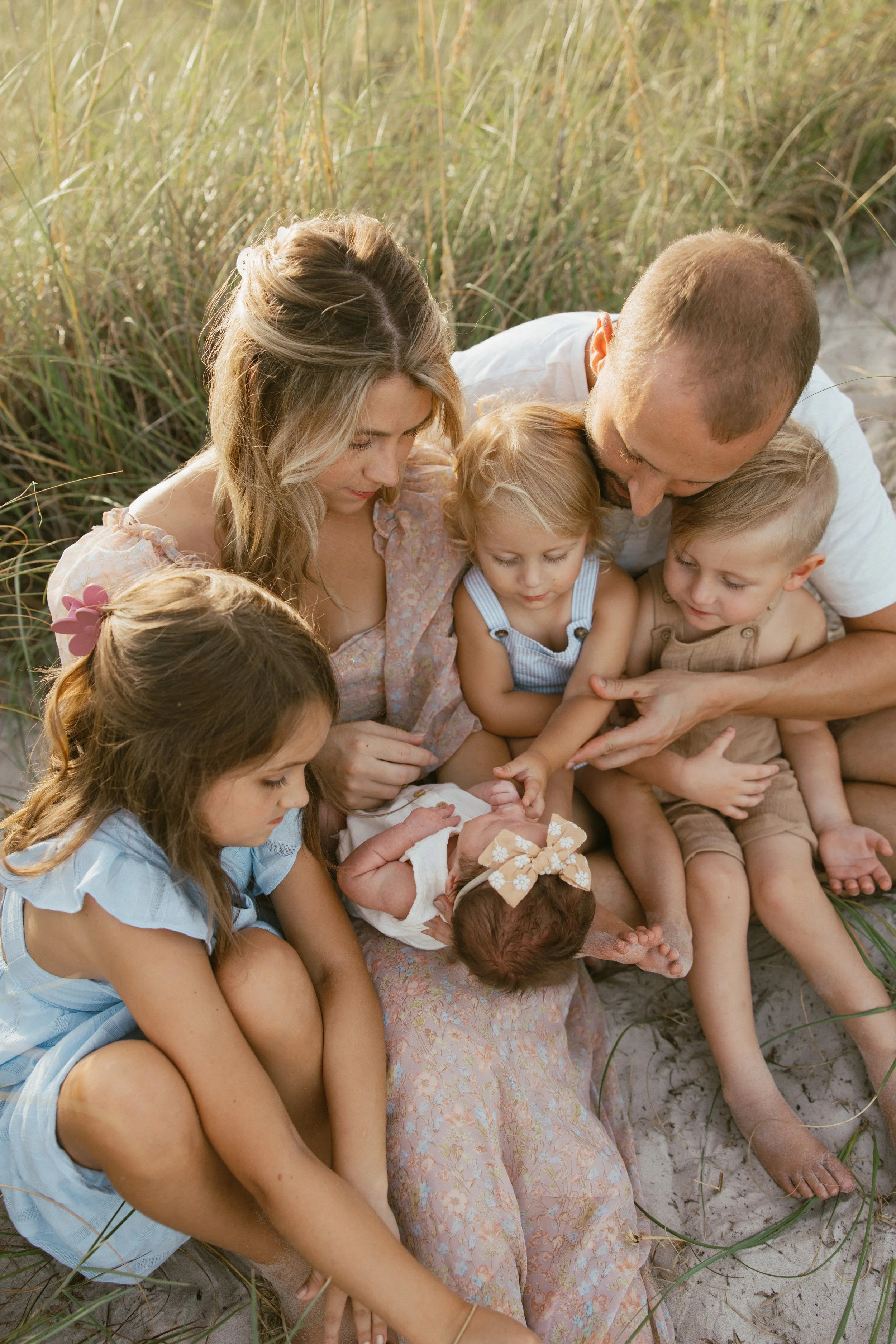 Family of six sitting on the sand in a grassy area, with a newborn baby at the center, surrounded by her parents and four young children, all engaged and looking at the baby.