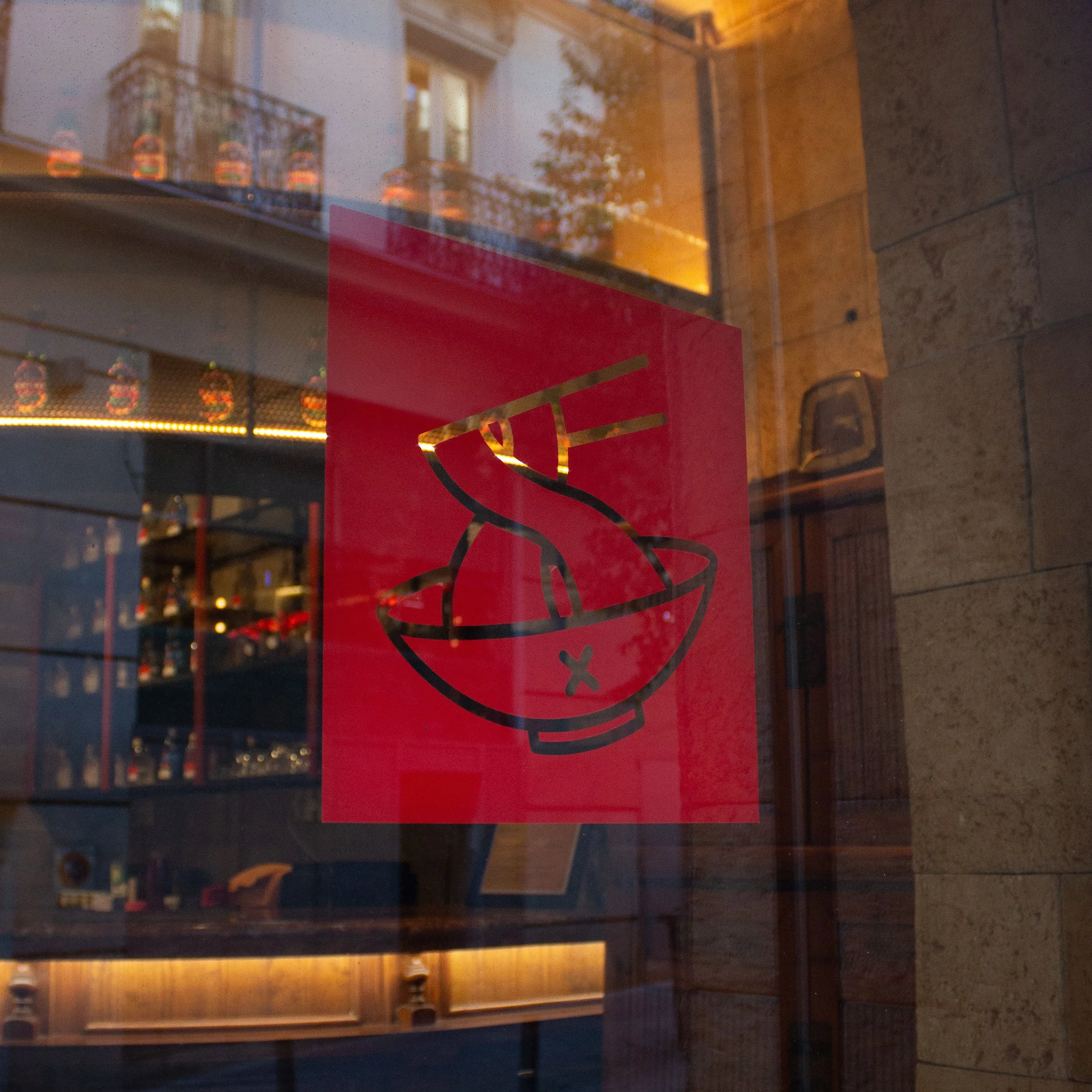 Imagen de una vitrina de tienda con las puertas de vidrio reflejando edificios y árboles del exterior, y un cartel rojo con diseño en negro.