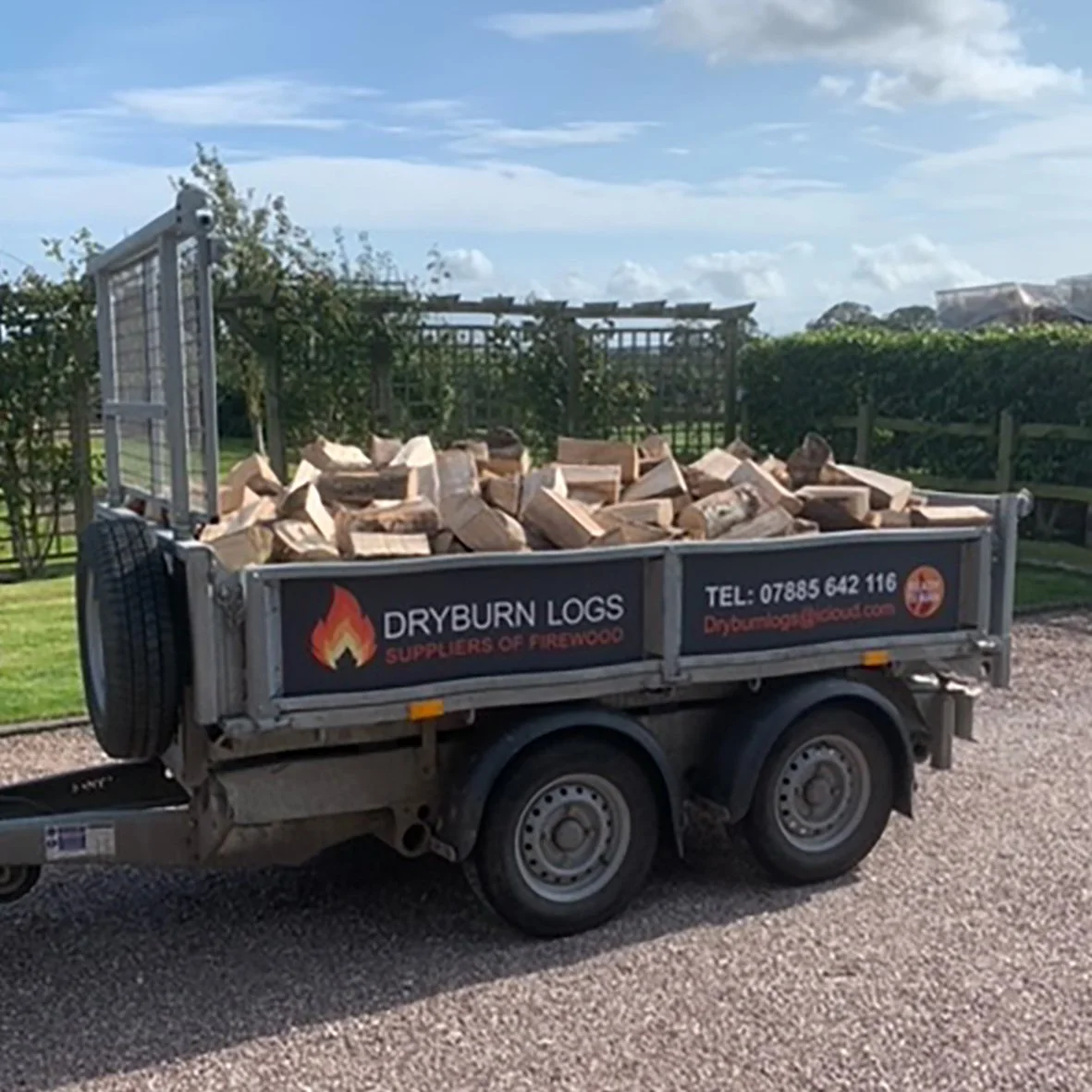 A trailer loaded with firewood from Dryburn Logs, parked on a gravel path. Trees and a wooden fence are visible in the background. The trailer bears a sign with contact details and imagery related to firewood supply.