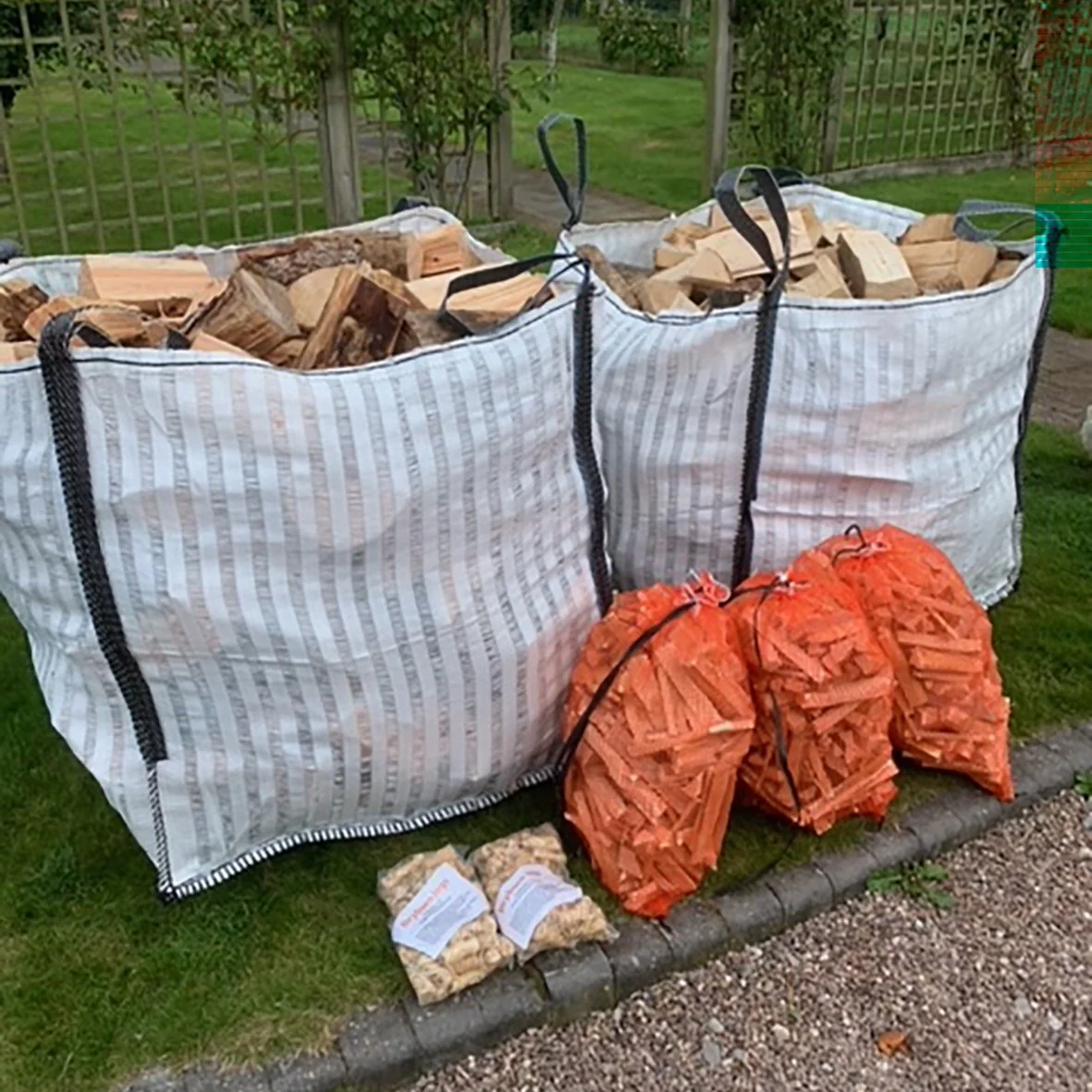 Two large bags of firewood labeled 'DRYBURN LOGS' are placed outside in a garden setting. The bags contain chopped logs and have contact information printed on them. Nearby are two green bags filled with kindling and a smaller bag of wood shavings, all arranged on a stone pathway.
