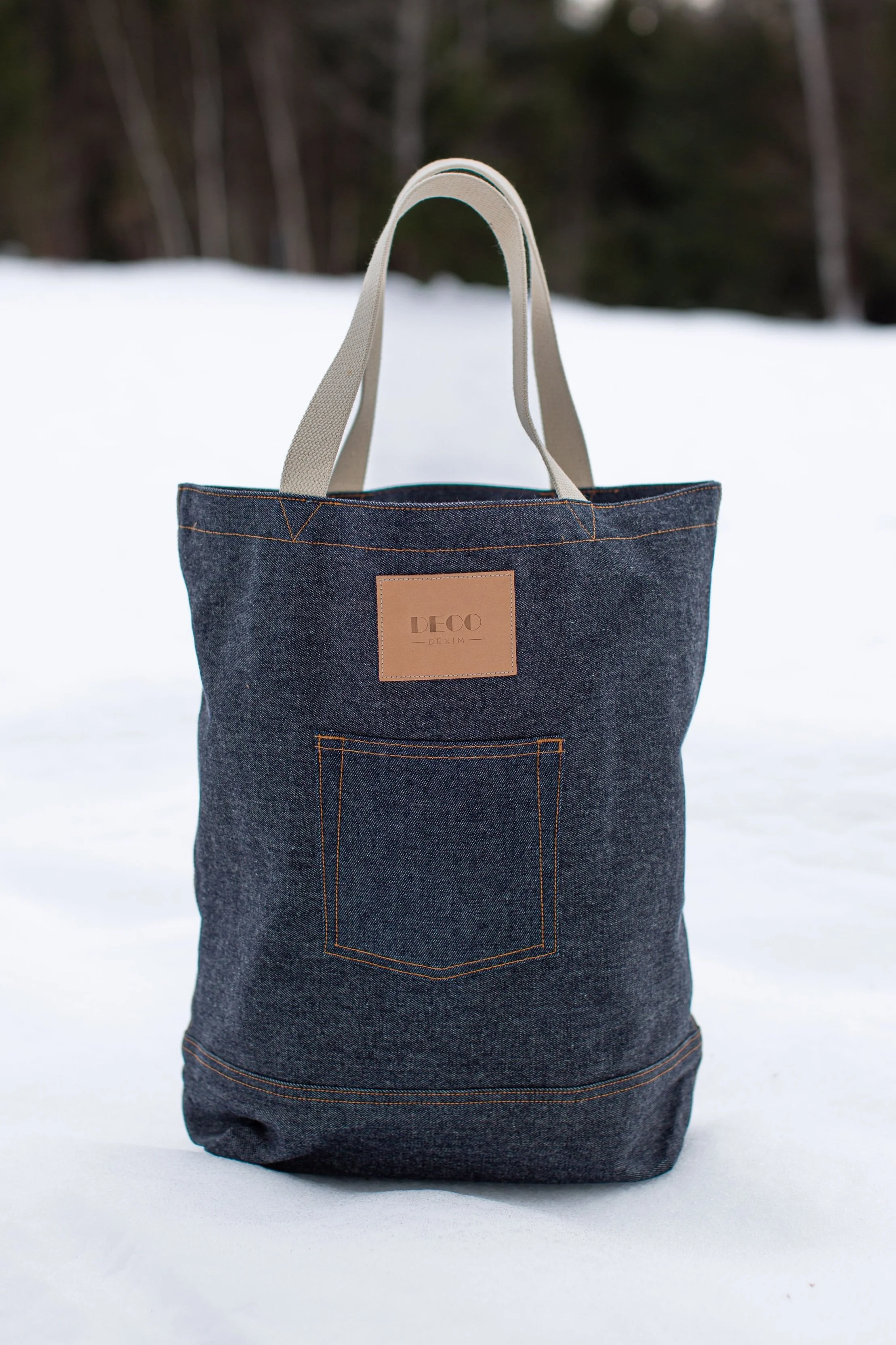 Denim tote bag with handles on snow, featuring a front pocket and a leather label.