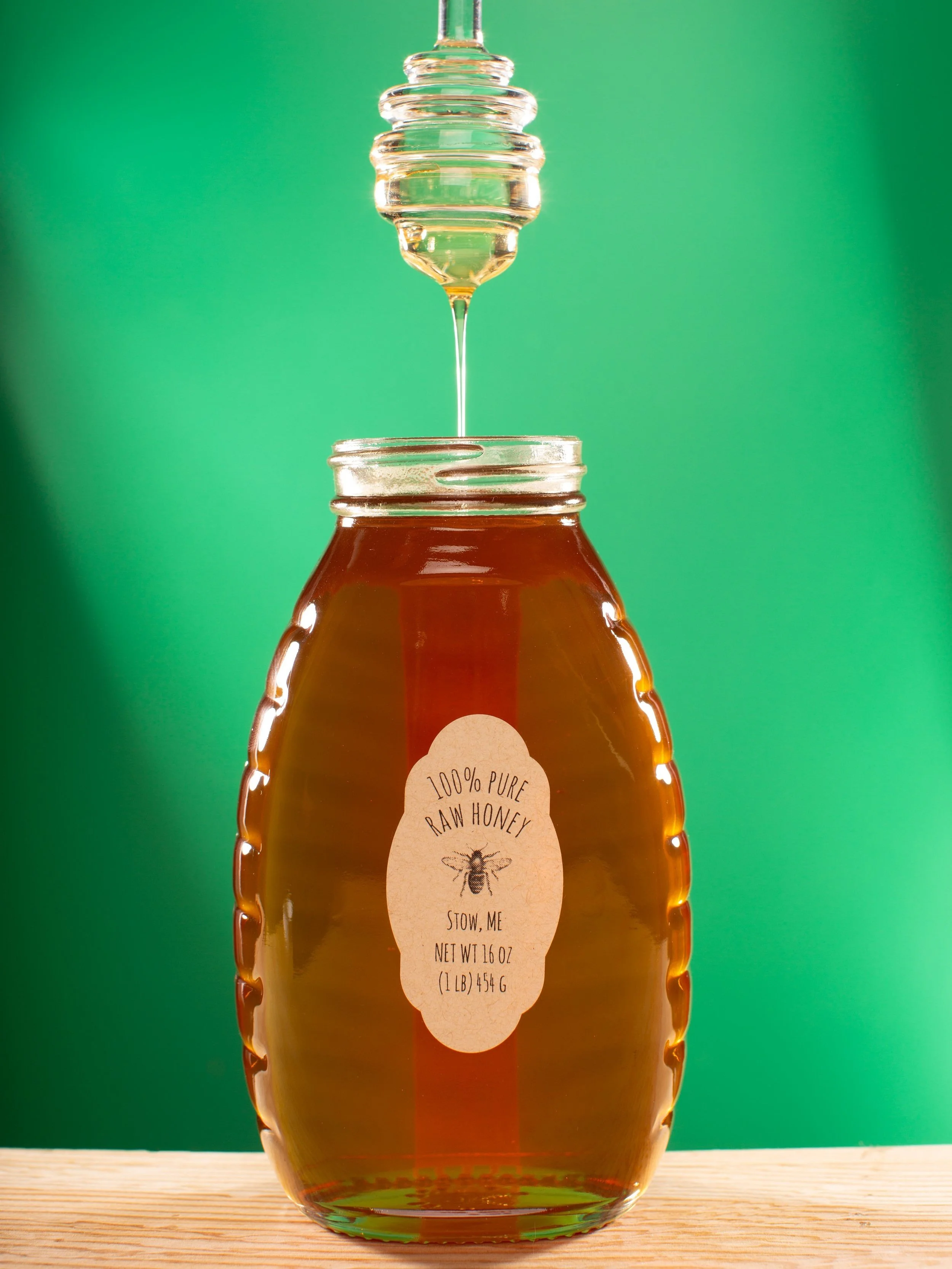 Jar of raw honey with a honey dipper drizzling honey into it, set against a green background.
