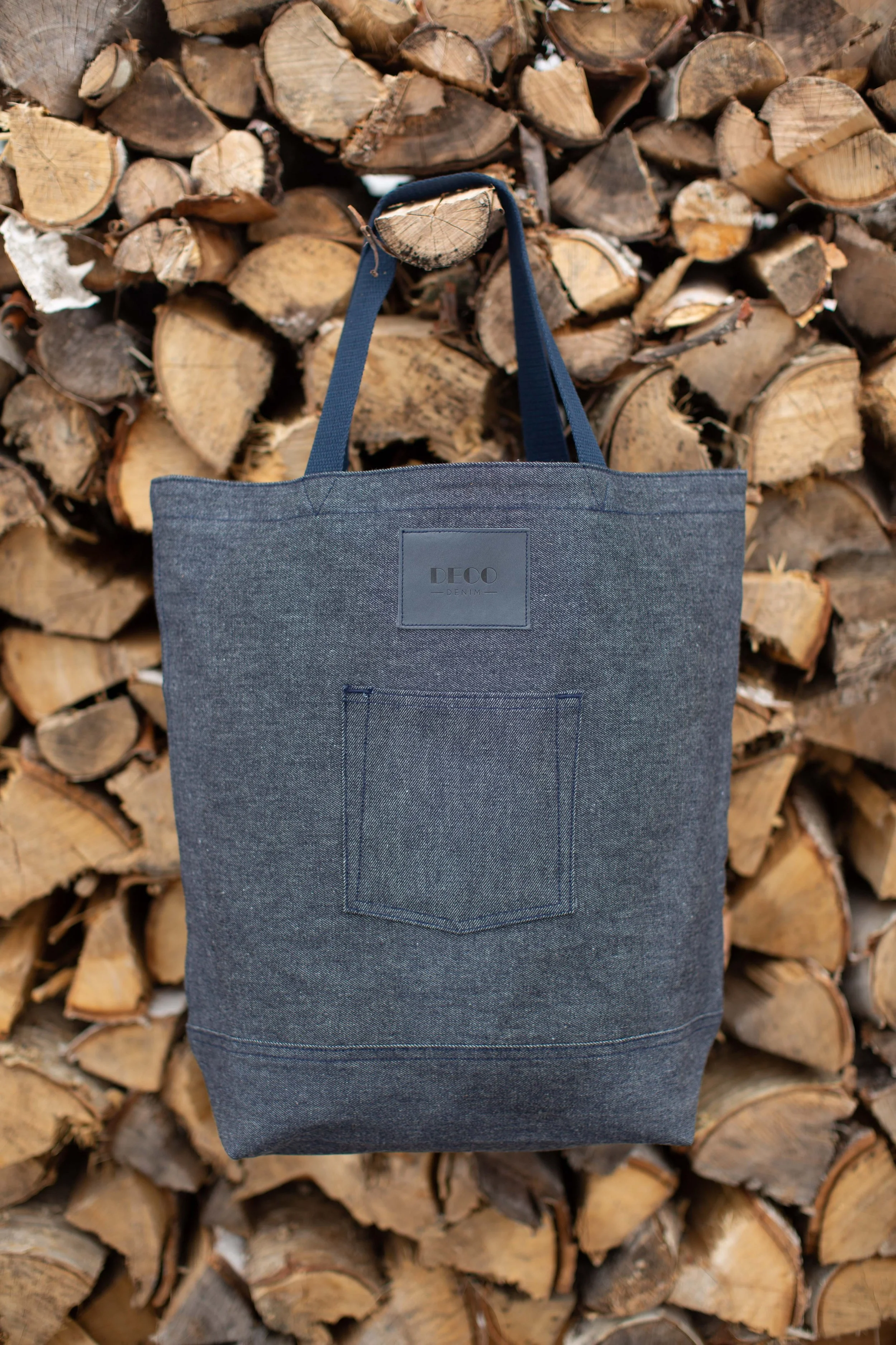 Blue denim tote bag with front pocket against stacked firewood background.