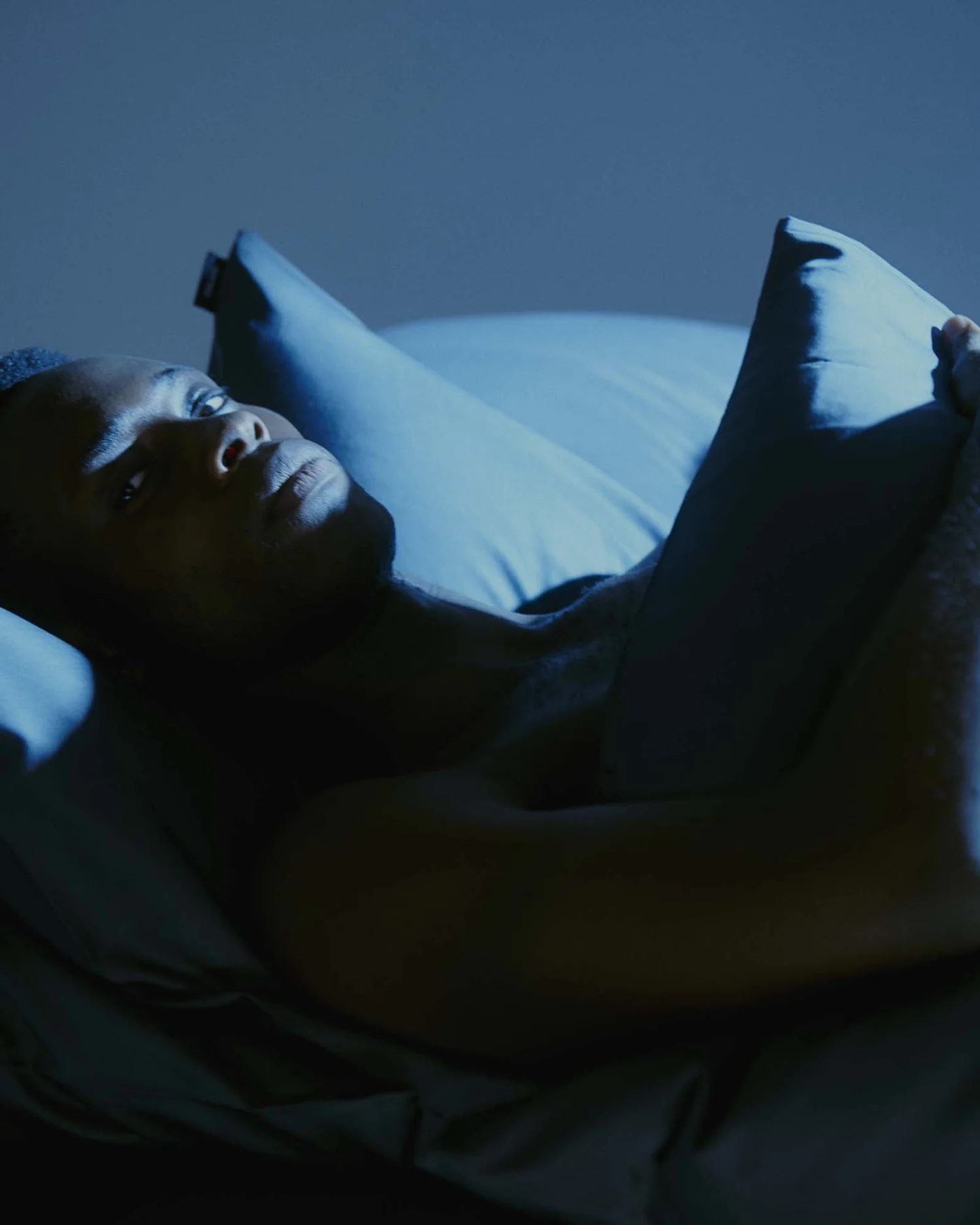 A model resting against pillows, looking towards the camera in deep blue light.