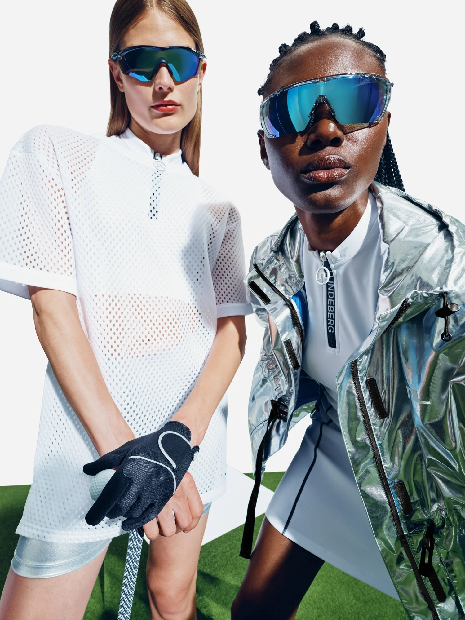 Two women dressed in sporty, futuristic golf attire wearing large mirrored sunglasses. They are standing on artificial green turf with a white background.