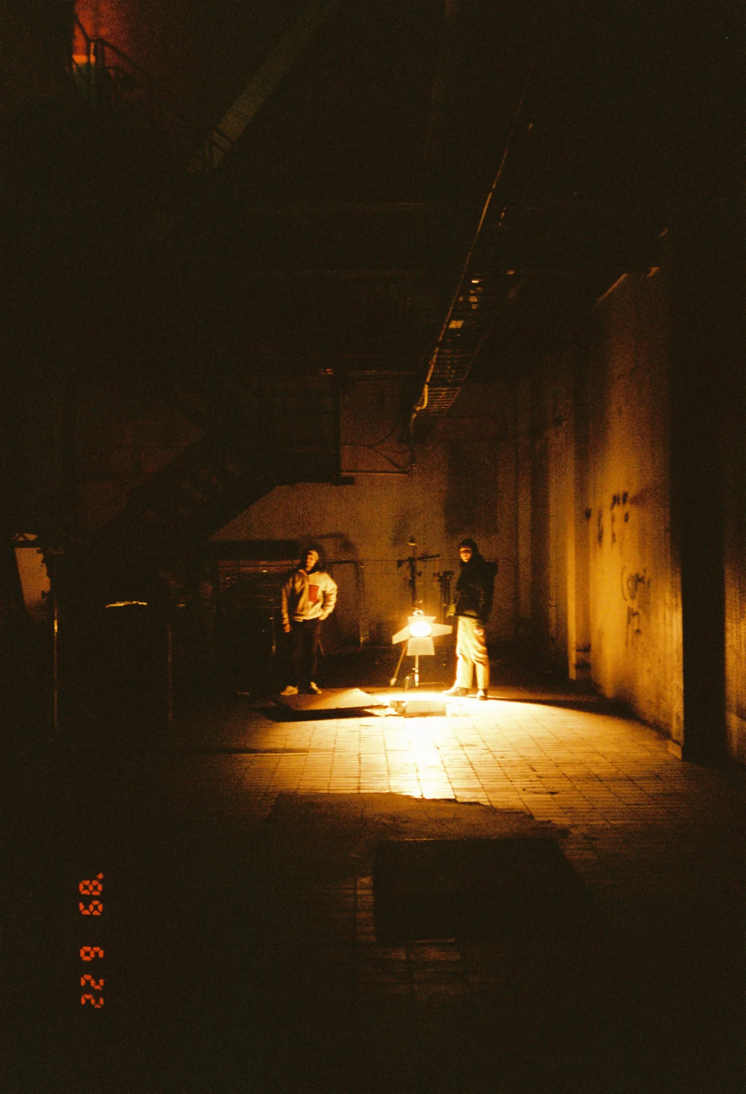 Two people stand in a dark, industrial space illuminated by a small bright lamp, with visible stairs and pipes overhead.