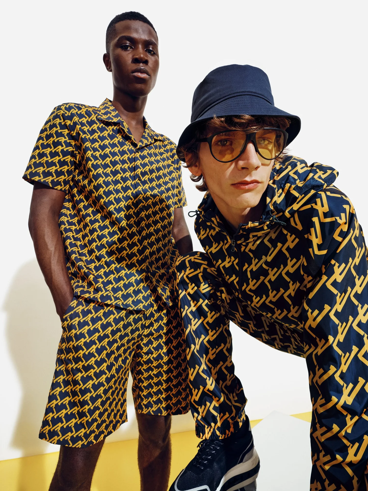 Two young men wearing matching navy and yellow geometric patterned outfits, one standing and the other crouching, against a white background with yellow accents.