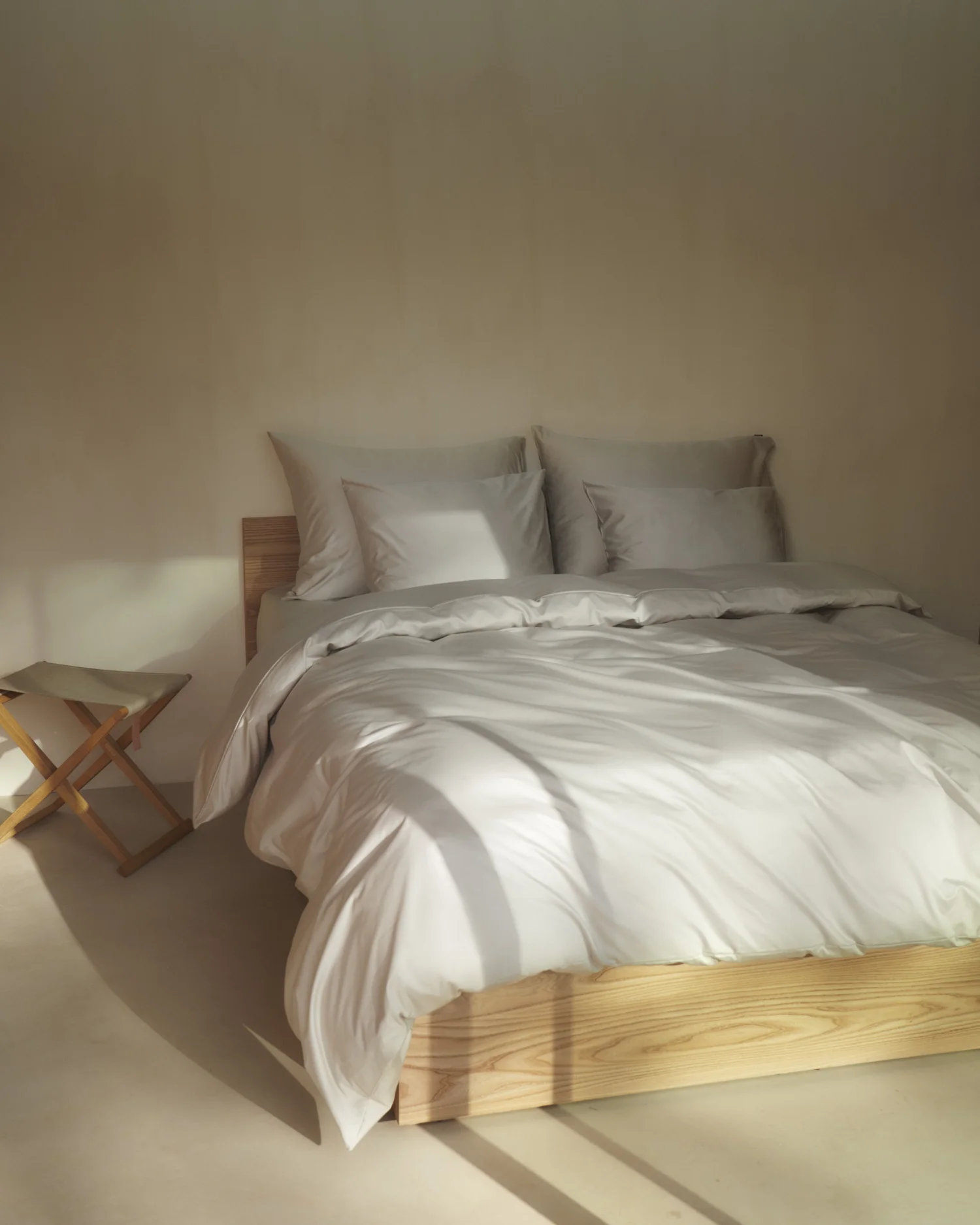 A neatly made bed with white bedding and four pillows, positioned against a beige wall. To the left, there's a small wooden stool with a cushioned top.