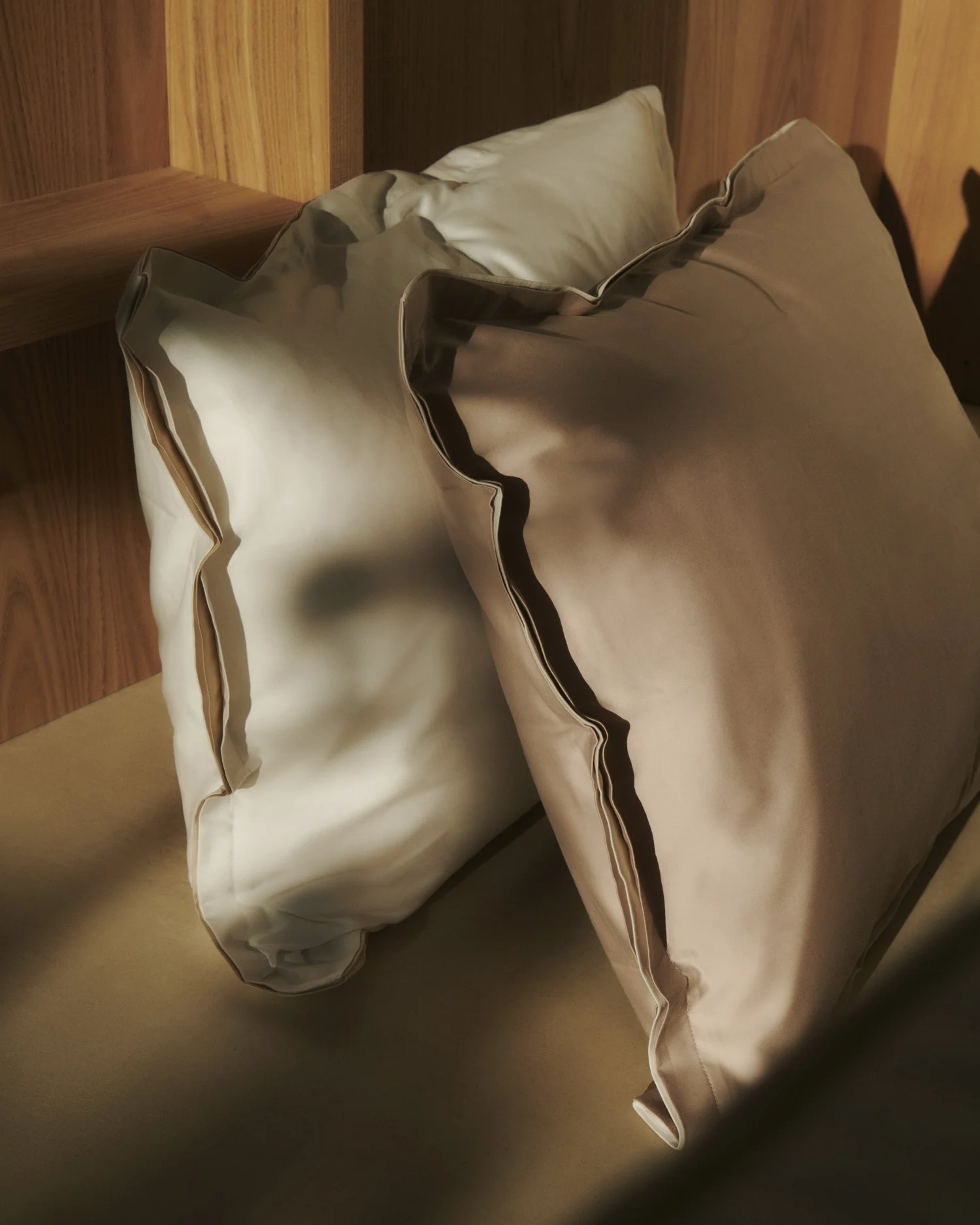 Two pillows with beige silk pillowcases and dark piping, resting on a bed with a wooden headboard.