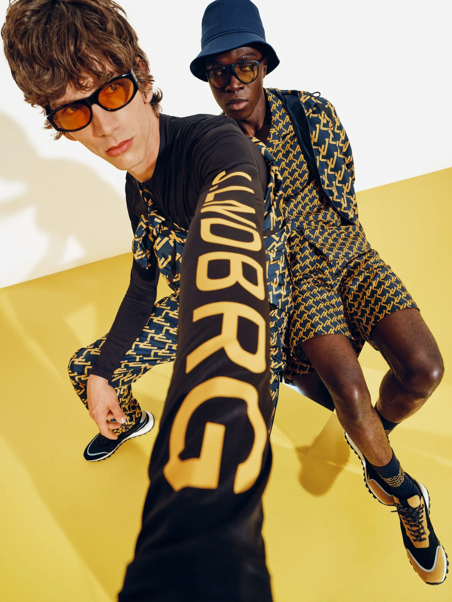 Two men in fashionable clothing and sunglasses pose for a photo on a yellow background.