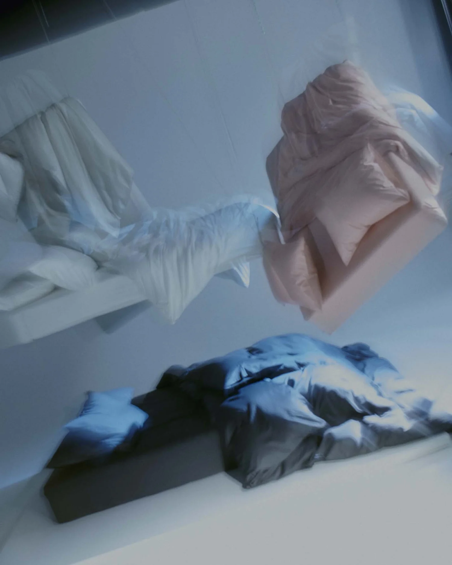 Upside-down view of crumpled bedsheets and pillows on a bed with a dark headboard, with blankets of various colors and a pink upholstered bedframe in the background.
