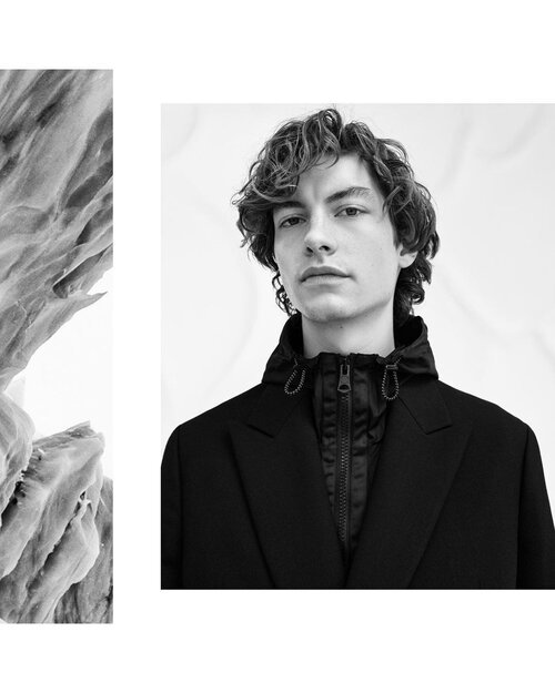 Black and white photo of a young man with curly hair wearing a black jacket standing against a white background.