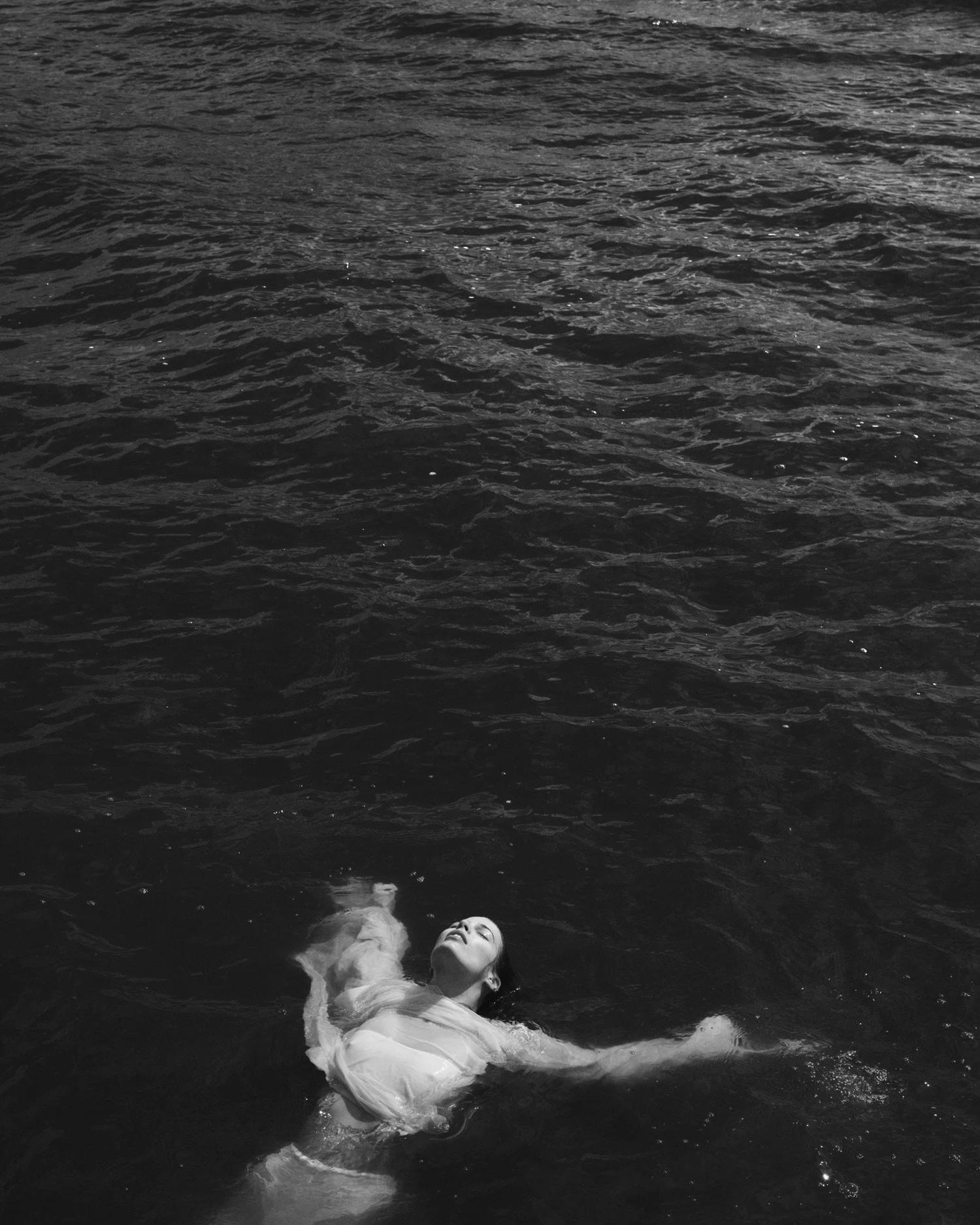 A woman in a white dress floating on her back in dark water with her head tilted back and eyes closed.