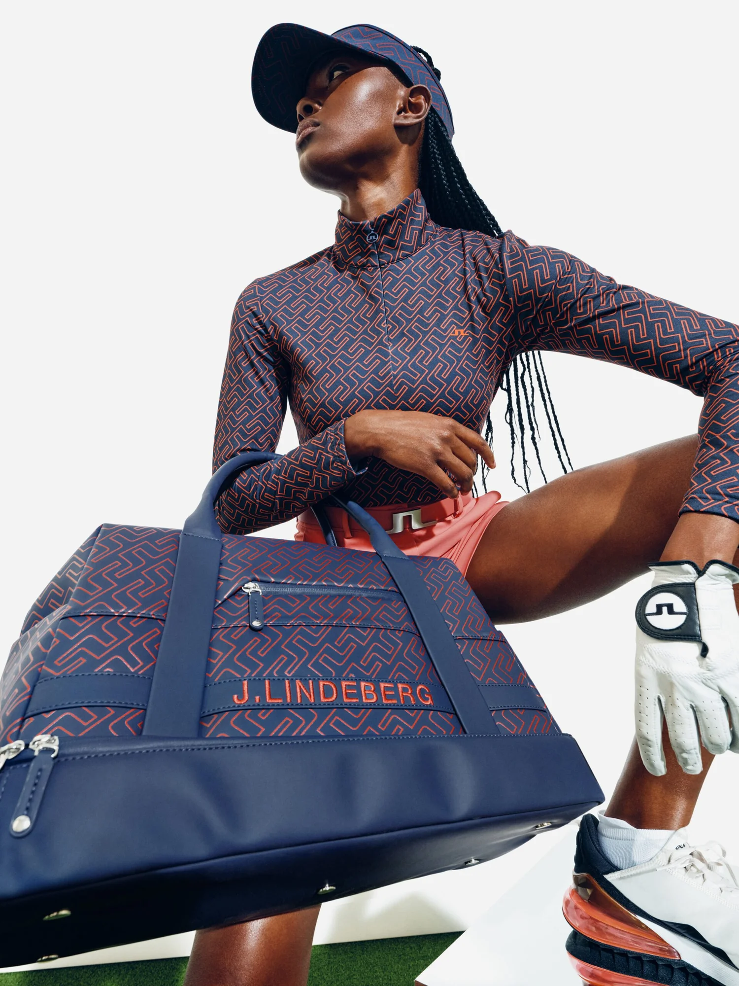 A woman dressed in athletic golf attire holding a navy blue, red, and gray golf bag with the brand name J. Lindeberg, standing in a pose with one hand on her hip and one leg bent, wearing a matching cap, patterned long-sleeve shirt, white golf glove,