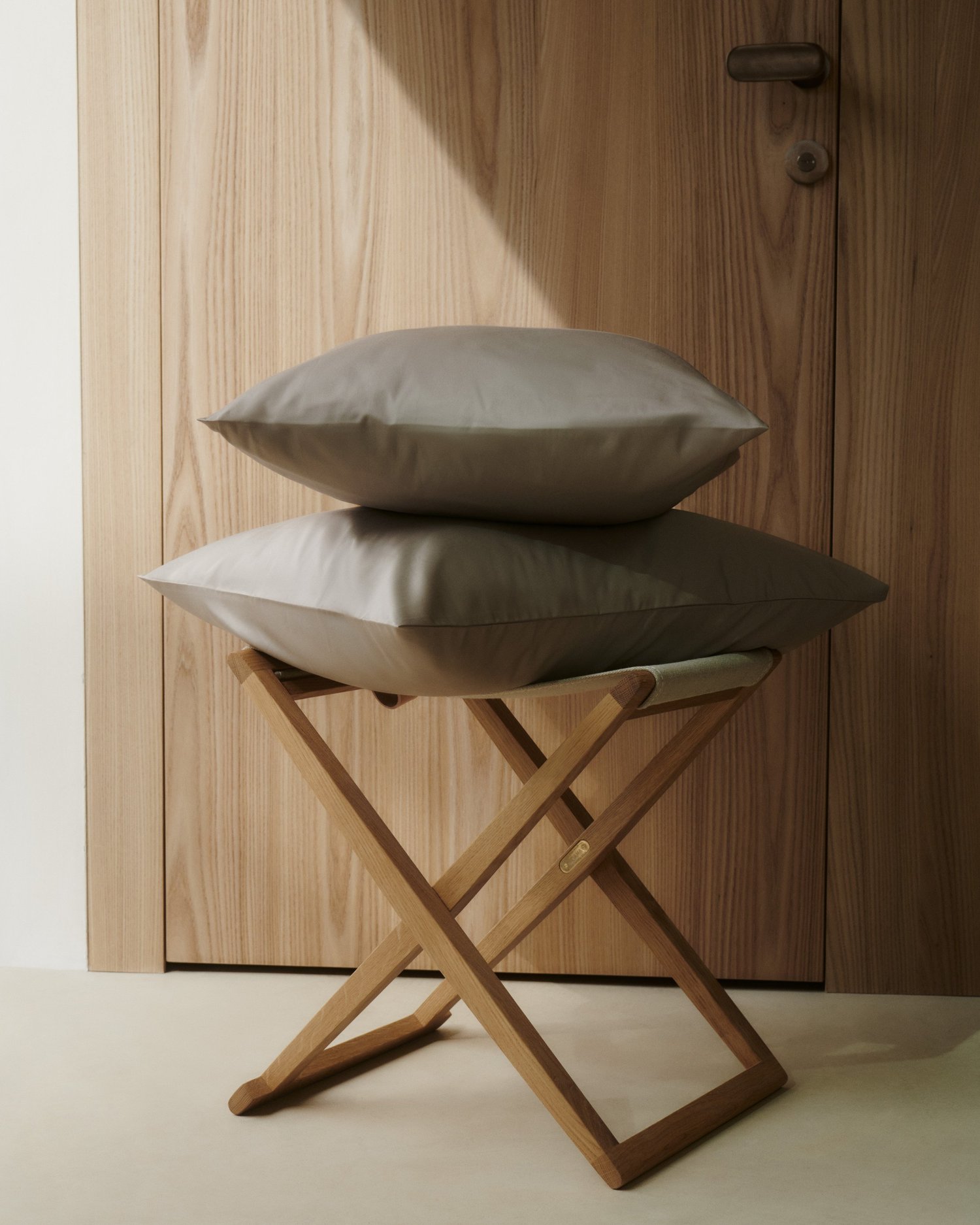Two beige pillows stacked on a wooden foldable stool in front of a wooden door.