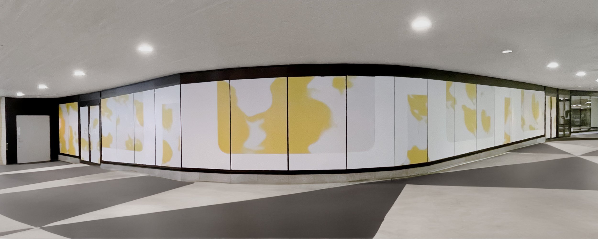 Empty hallway with a curved wall decorated with large abstract yellow and white artwork, gray and white patterned floor, ceiling with recessed lighting, and glass doors at the far end.