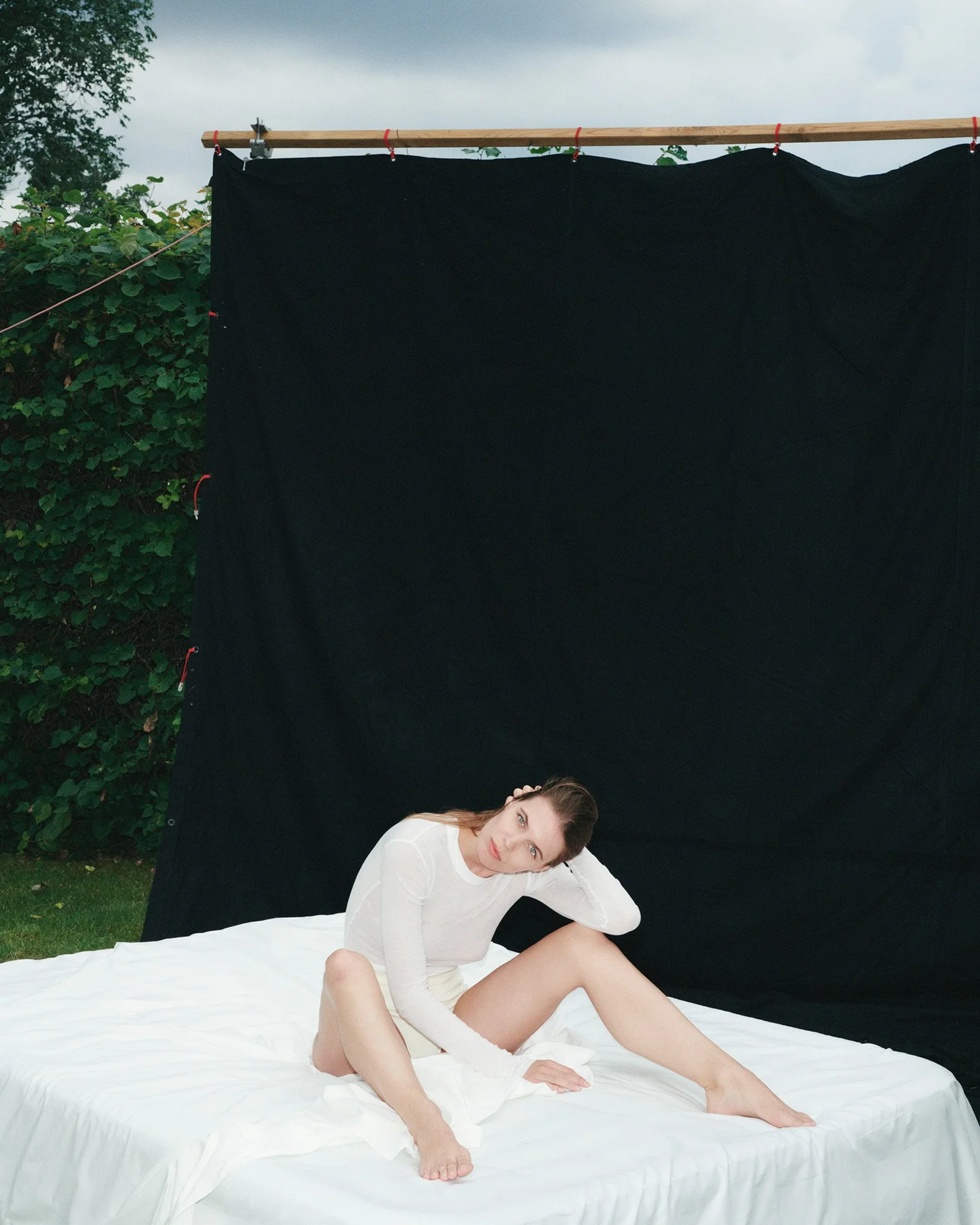 A woman in a white long-sleeve top and shorts sitting on a bed outdoors with a black backdrop behind her, and greenery and cloudy sky in the background.
