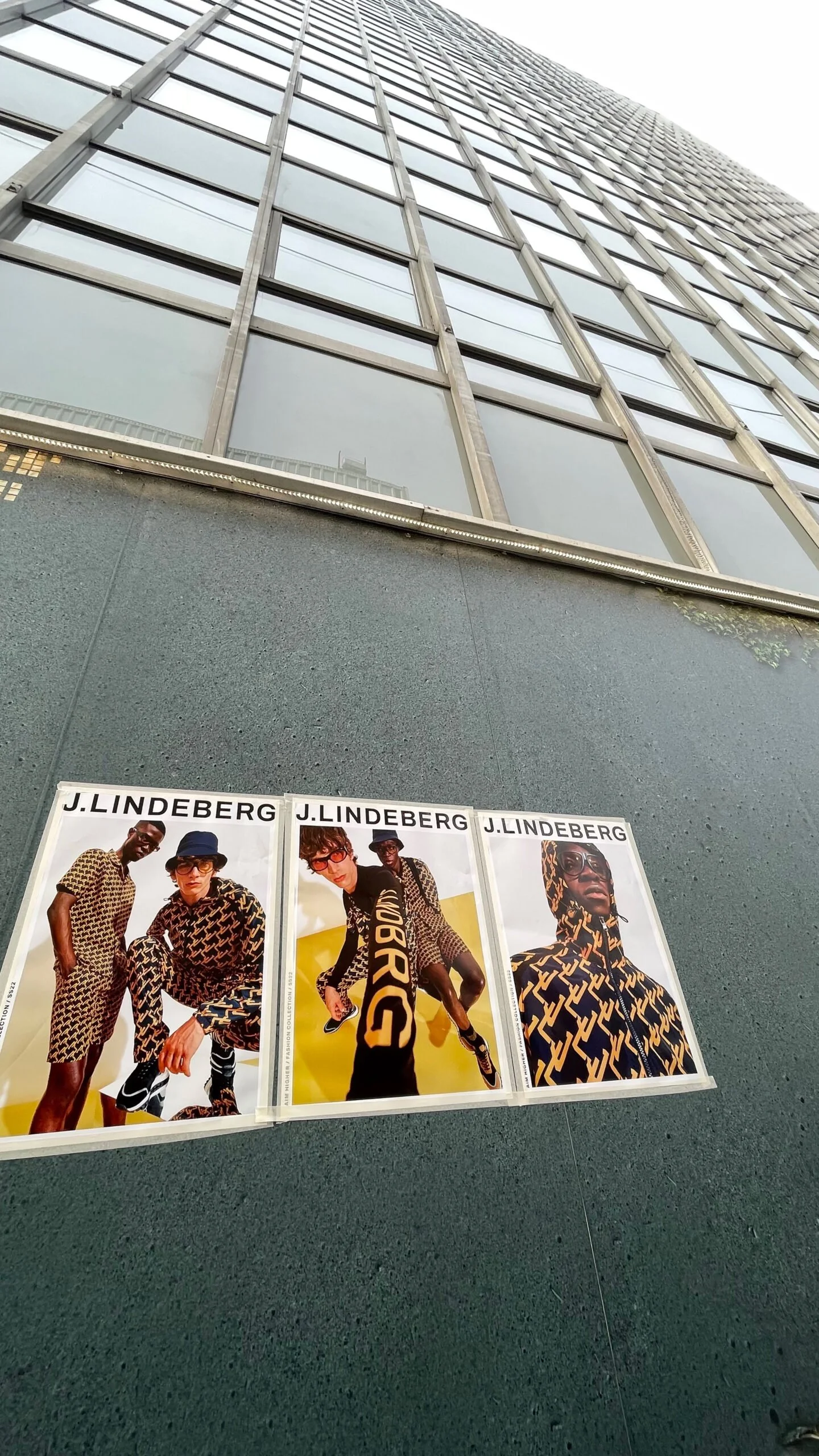 Three posters for J. Lindeberg fashion brand on a gray wall outside a tall glass building, showing models wearing designer clothing