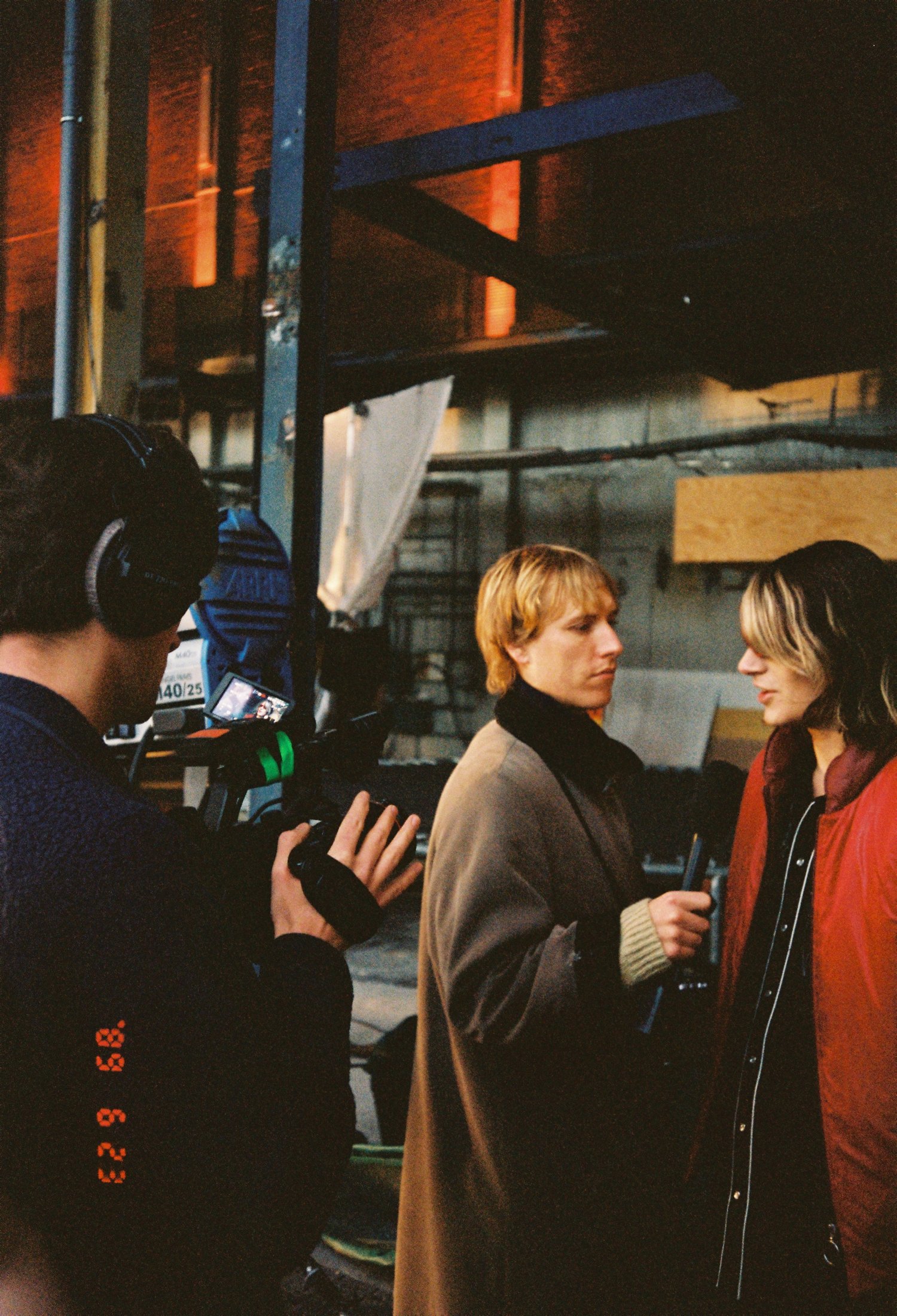 Two women being filmed in a warehouse or studio while a man operates a professional video camera. One woman has short blond hair and wearing a brown blazer; the other has longer hair with highlights and is wearing a red jacket.