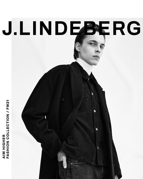 Black and white fashion advertisement featuring a young woman wearing a black jacket and shirt, with the name J. Lindeberg at the top and text indicating Aim Higher Fashion Collection.
