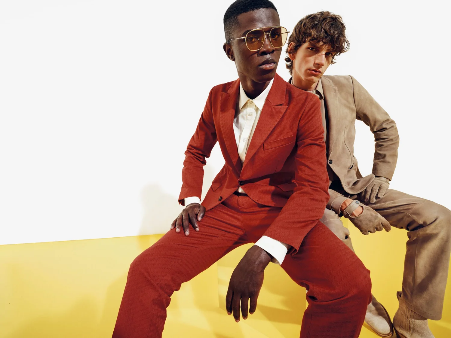 Two men in colorful suits sitting on a yellow surface against a plain white background. The man in front is wearing a red suit with a white shirt and glasses, while the man behind is in a beige suit with matching gloves and sneakers.
