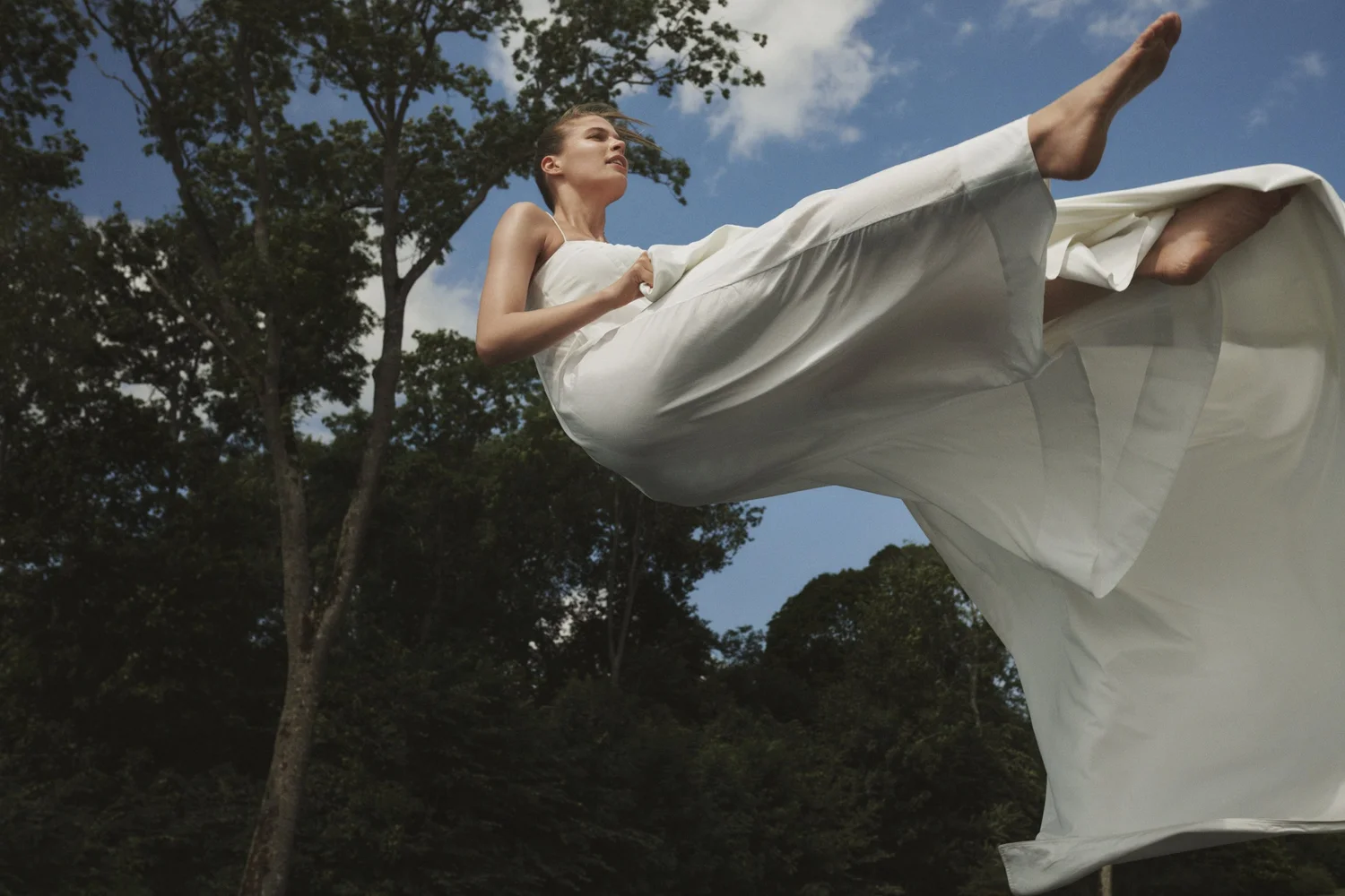 A low-angle view of a model lifting a white sheet against blue sky and trees, from the Ogland SS21 campaign.