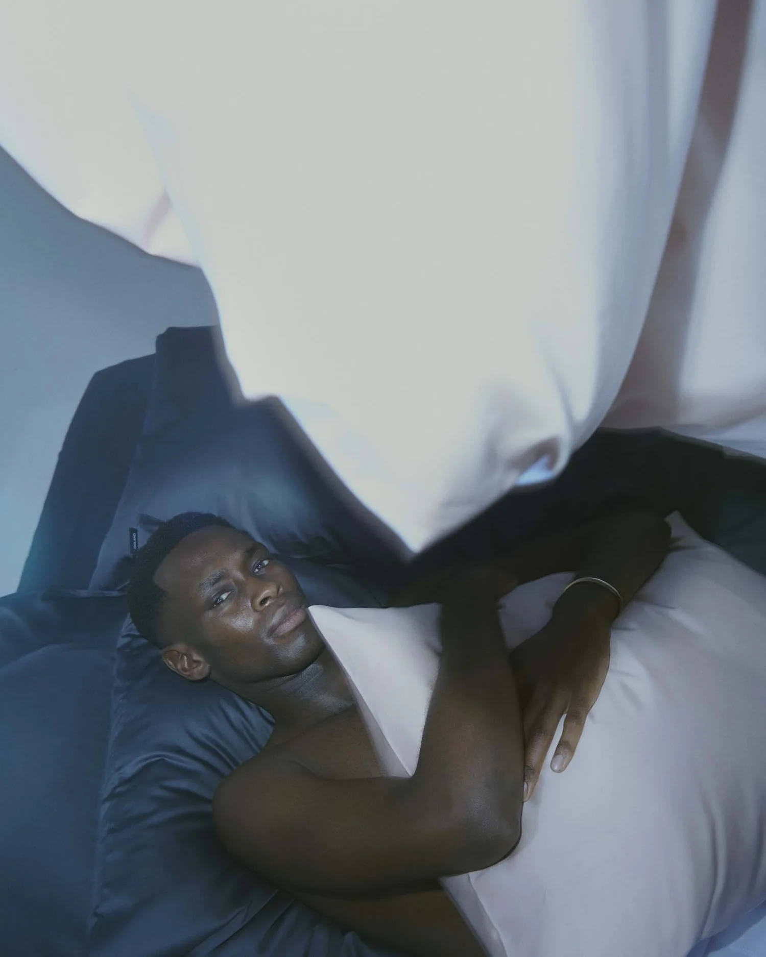 A man lying on a bed, looking at the camera with a serious expression, resting on one arm, with a white pillow and dark bedding.