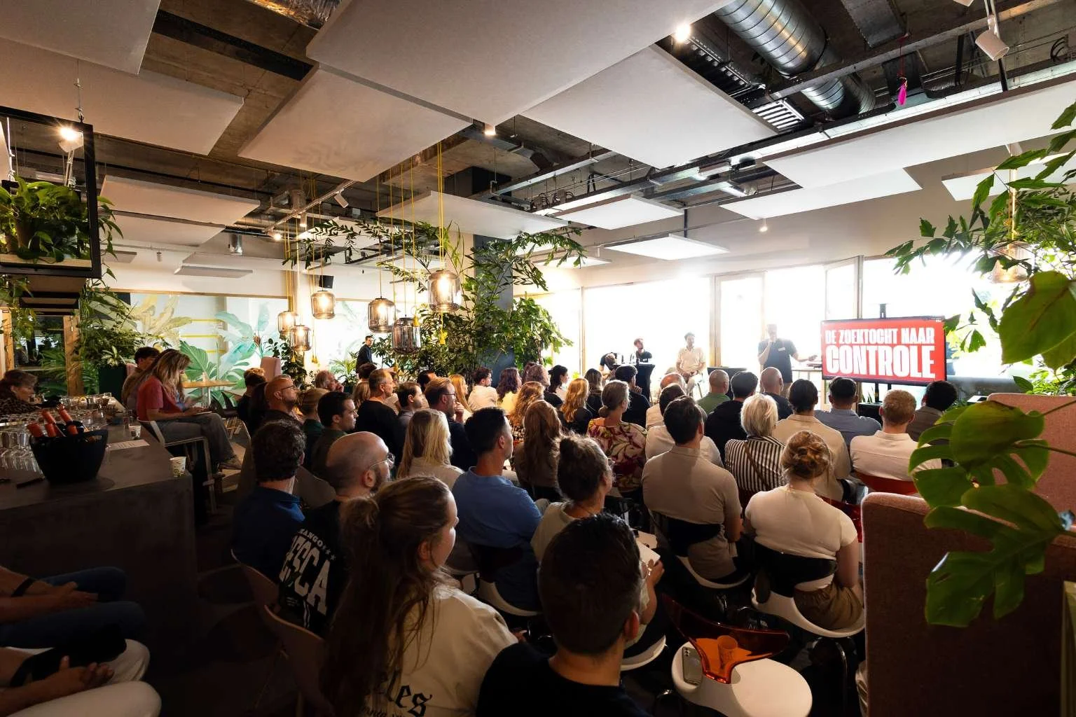 Grote groep mensen bij conferentie of presentatie in een modern, licht interieur met planten en grote ramen, met een scherm waarop 'De Zorgenlijst Naar Controle' staat.
