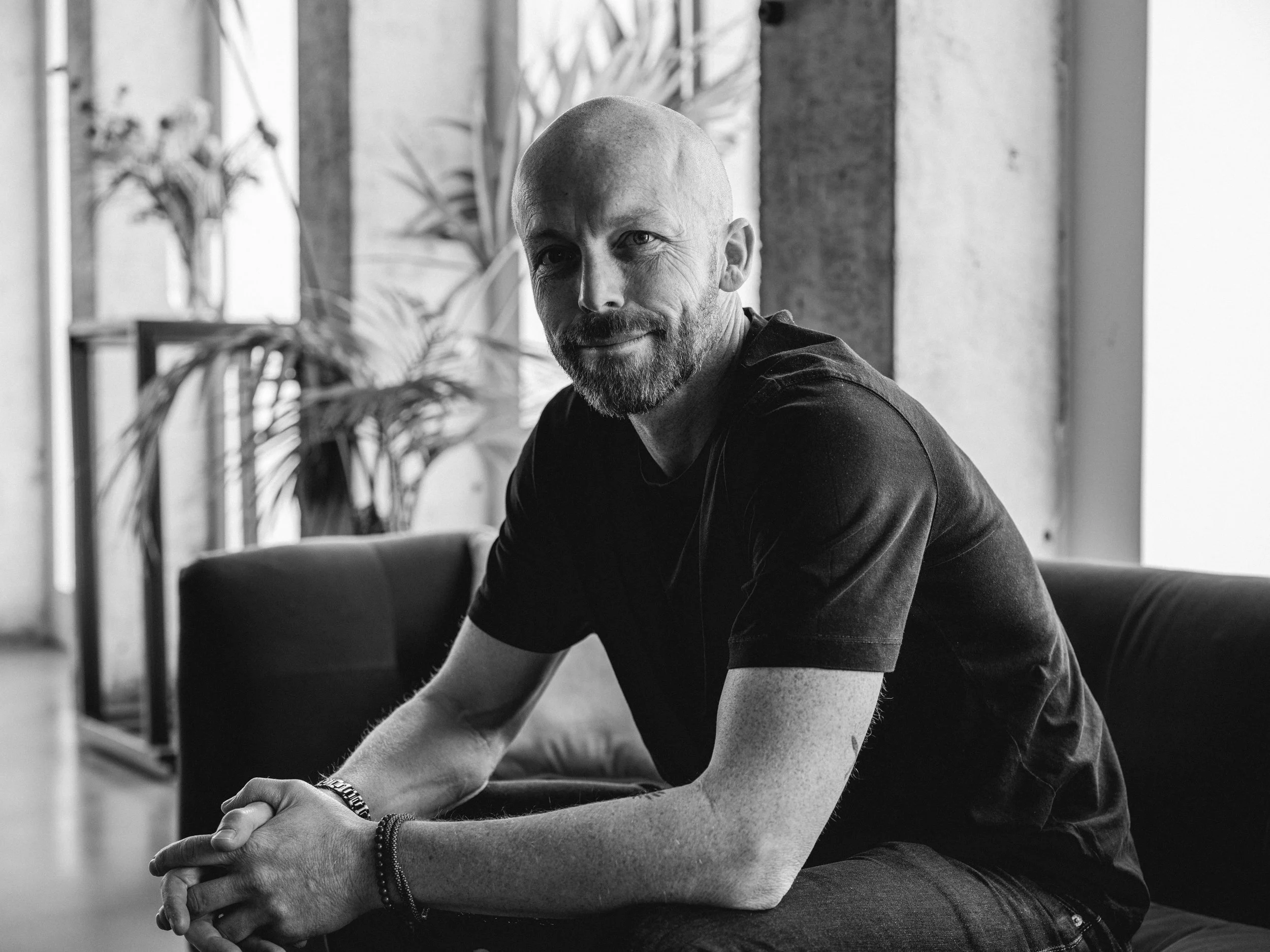 Zwart-wit foto van een man met een baard, zittend op een bank, lachend, met planten en ramen op de achtergrond.