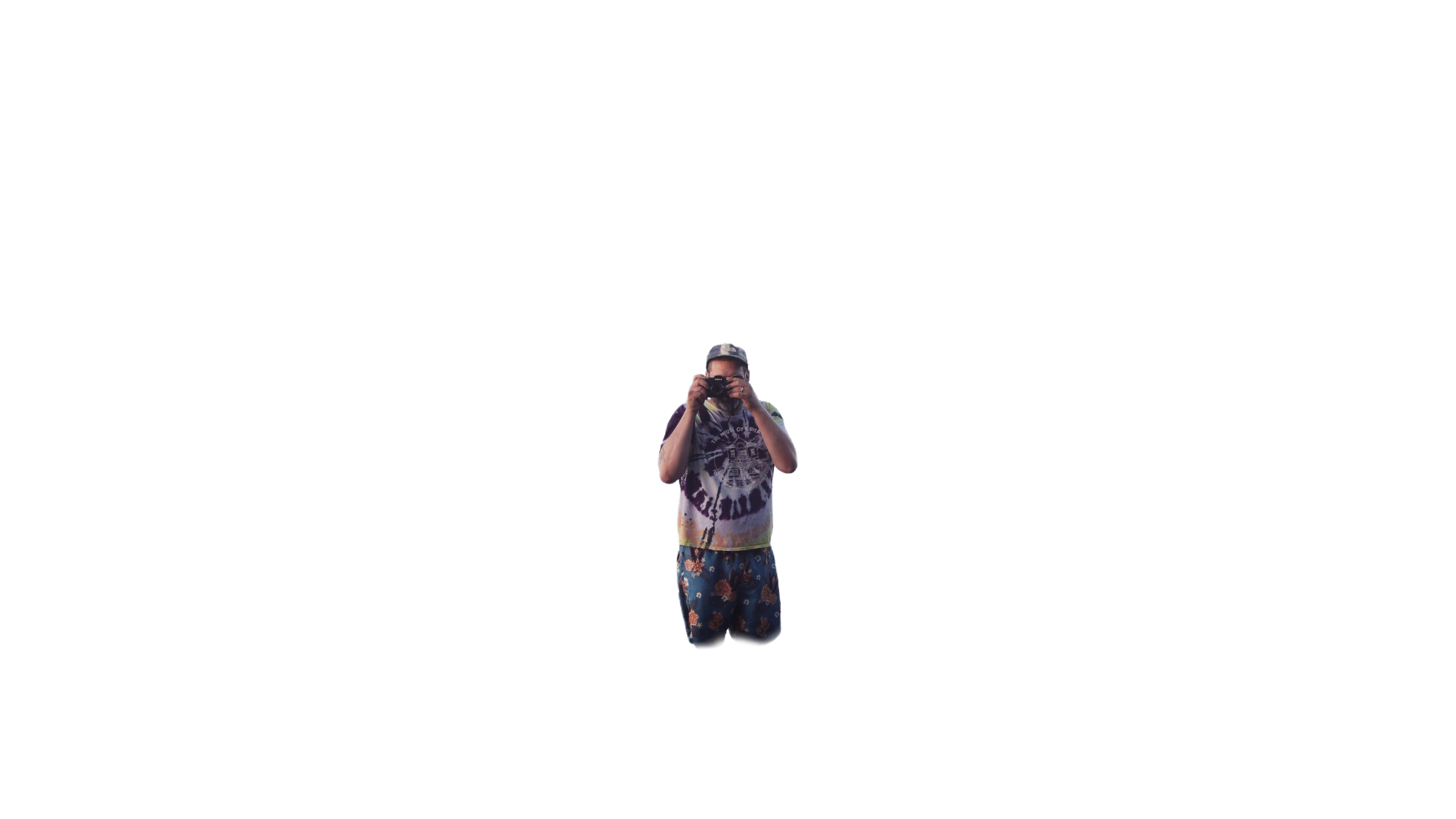 A person wearing a hat, glasses, a tie-dye shirt, and floral shorts, standing outdoors and taking a photo with a camera.