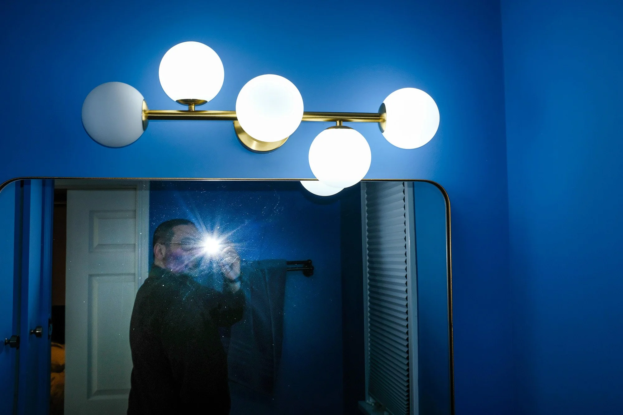 A person taking a selfie in a mirror with a flash on, showing a modern wall-mounted light fixture with four spherical white globes on a brass horizontal bar against a blue wall.