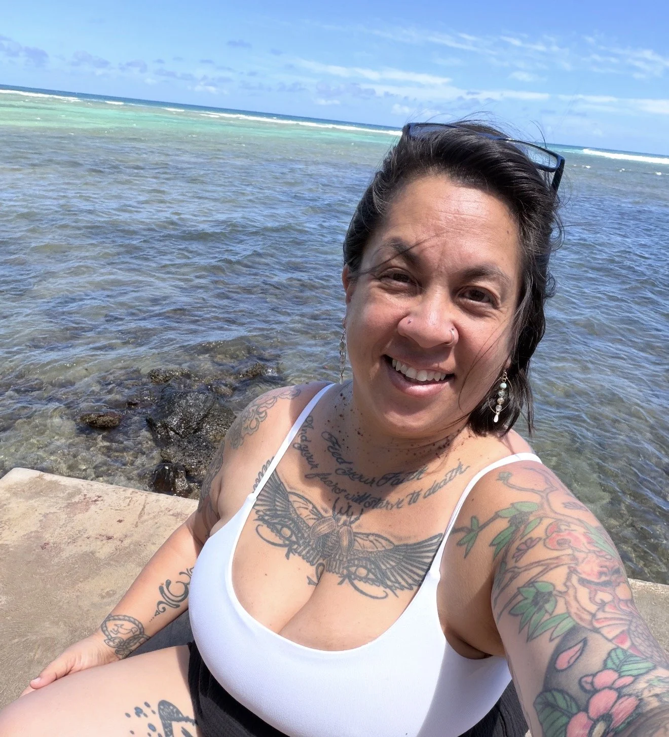 A woman with tattoos and earrings taking a selfie on a beach with clear water and blue sky in the background.