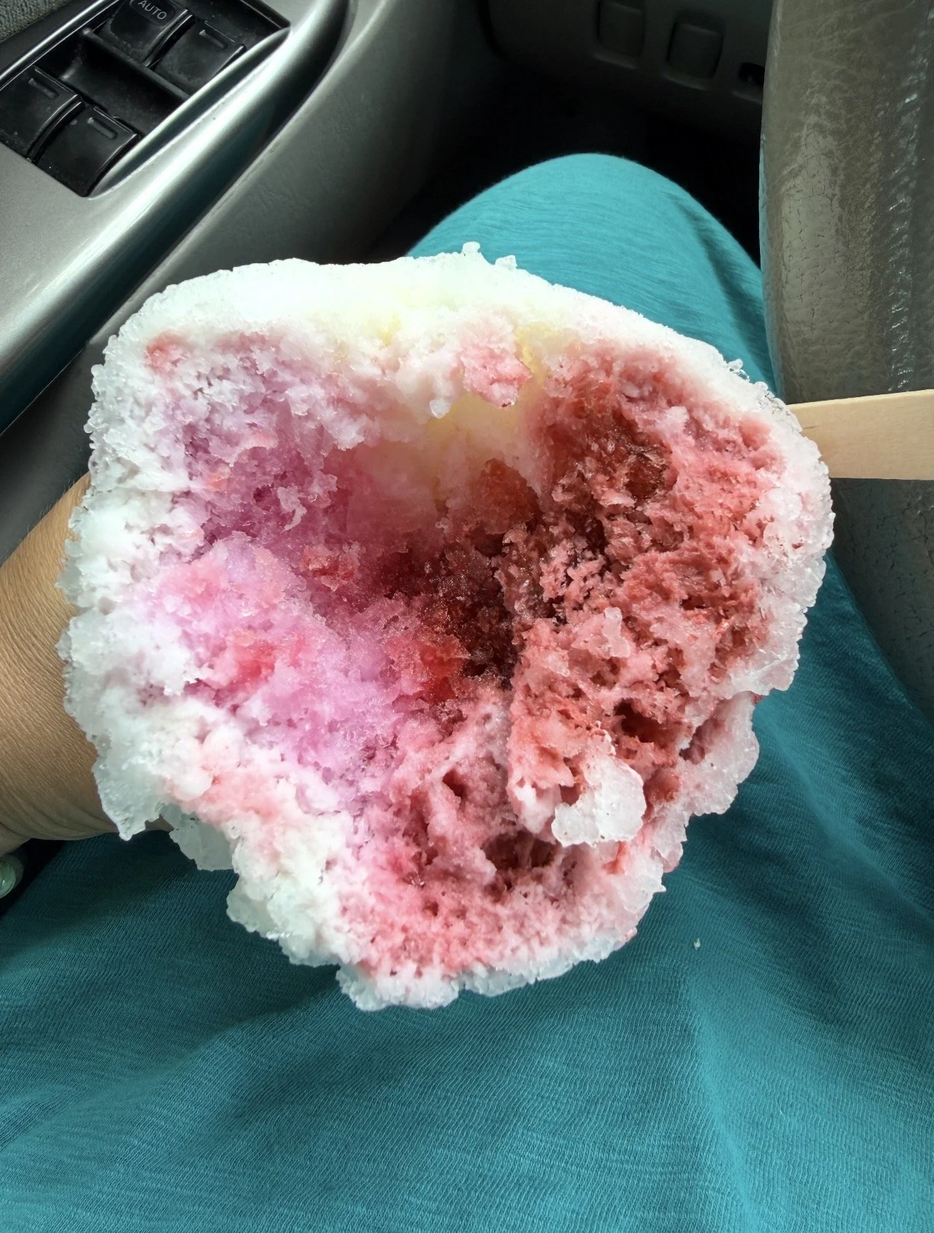 Close-up of a partially eaten snow cone with pink syrup on white ice, held by a person wearing teal pants, sitting inside a vehicle.